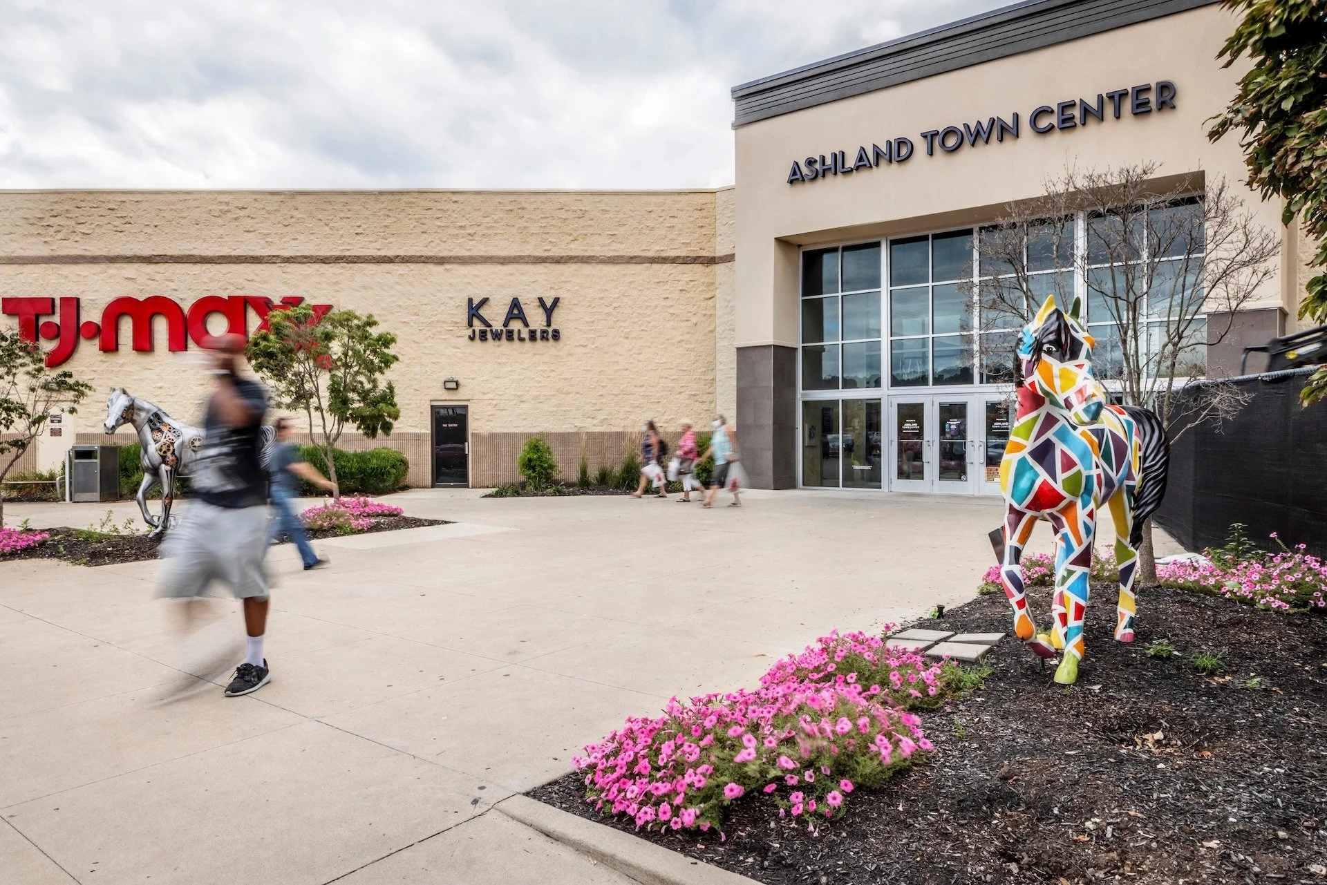 People walking outside Ashland Town Center shopping mall with colorful horse statues and pink flowers in the foreground.