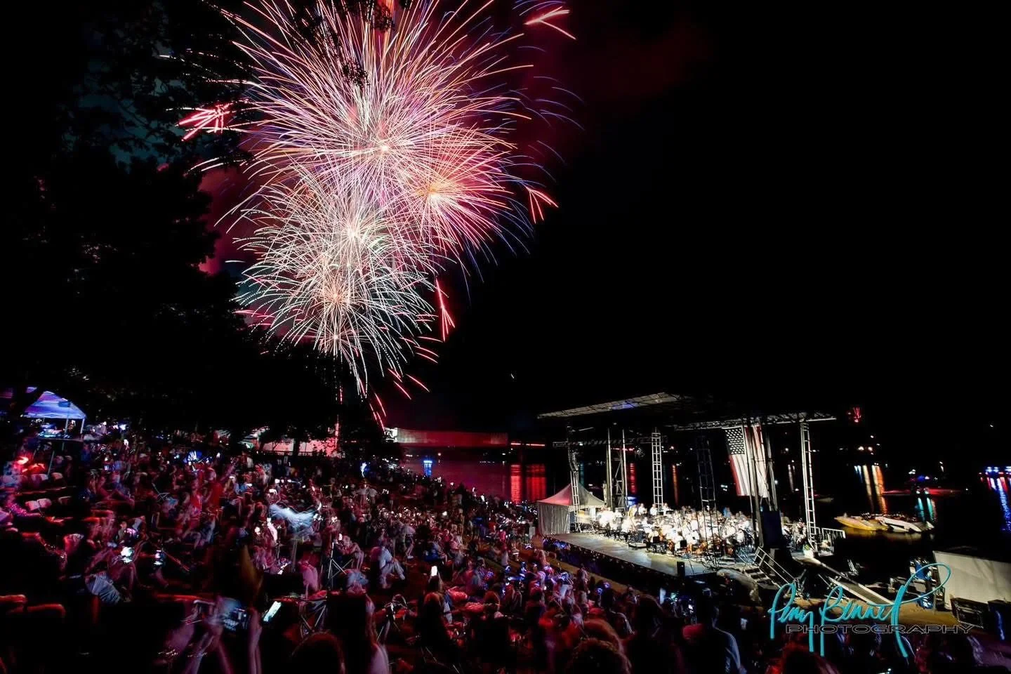 Fireworks display over an outdoor concert with a large audience seated watching a musical performance at night.