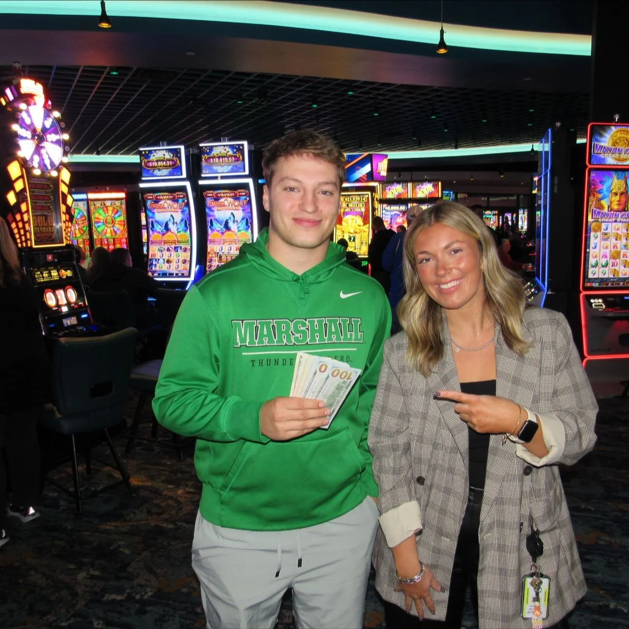 A young man wearing a green Nike hoodie and khaki pants holds euro bills, standing next to a smiling woman in a plaid blazer pointing at his money inside a brightly lit casino with slot machines in the background.
