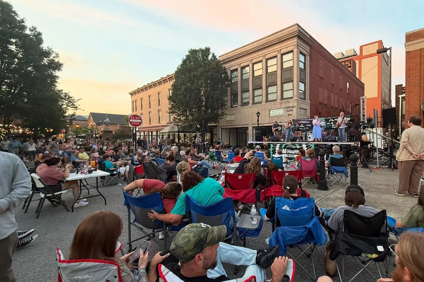 Outdoor concert in a town square with people sitting on chairs and enjoying a live band on a stage, with buildings and trees in the background during sunset.