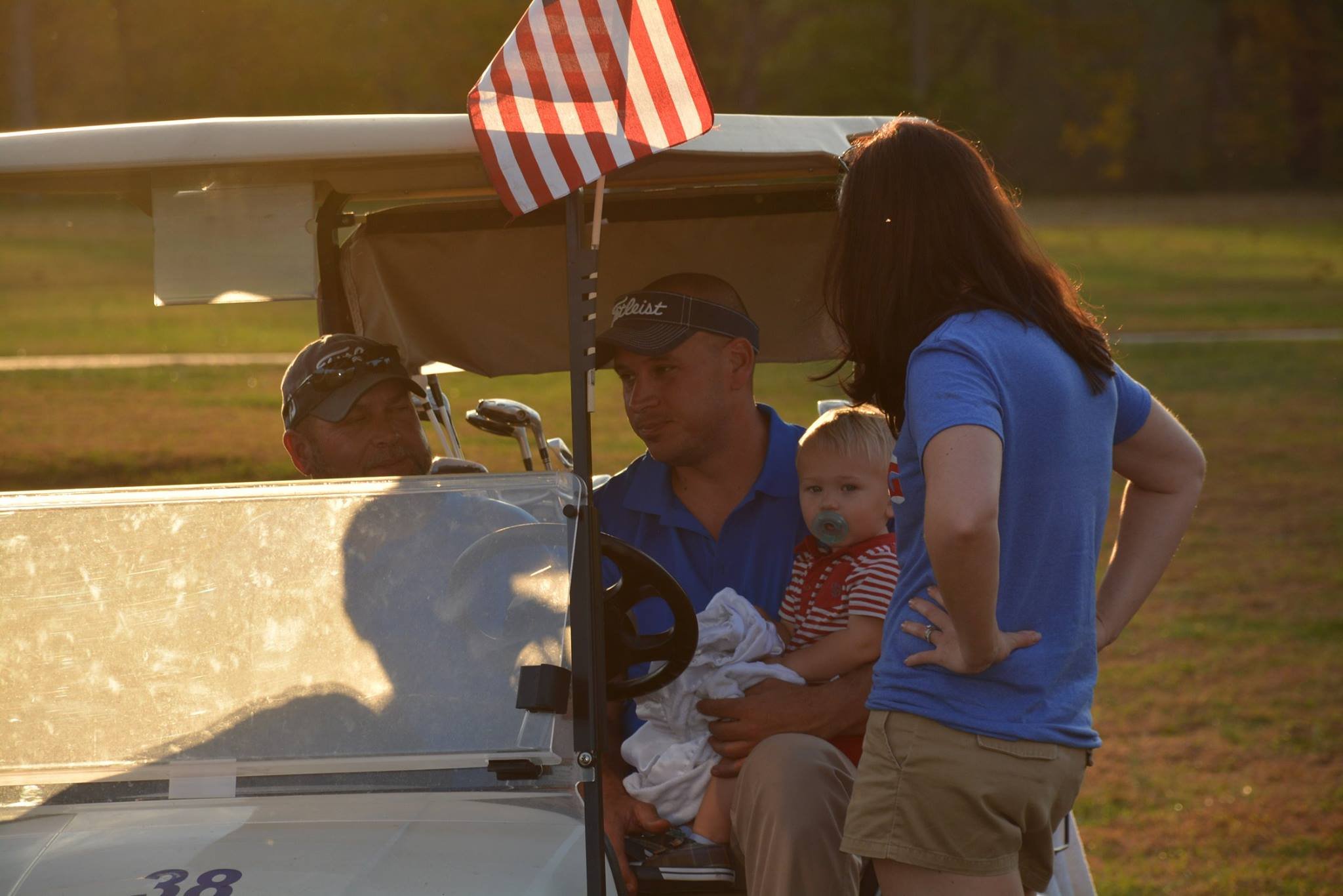 A woman in a blue shirt stands next to a man in a blue polo shirt sitting in a golf cart. The man is holding a young boy with a pacifier, wearing a red and white striped shirt. The woman and the man are engaged in conversation while the boy looks ahead. The golf cart has an American flag attached and the background is outdoors on a grassy field at sunset.