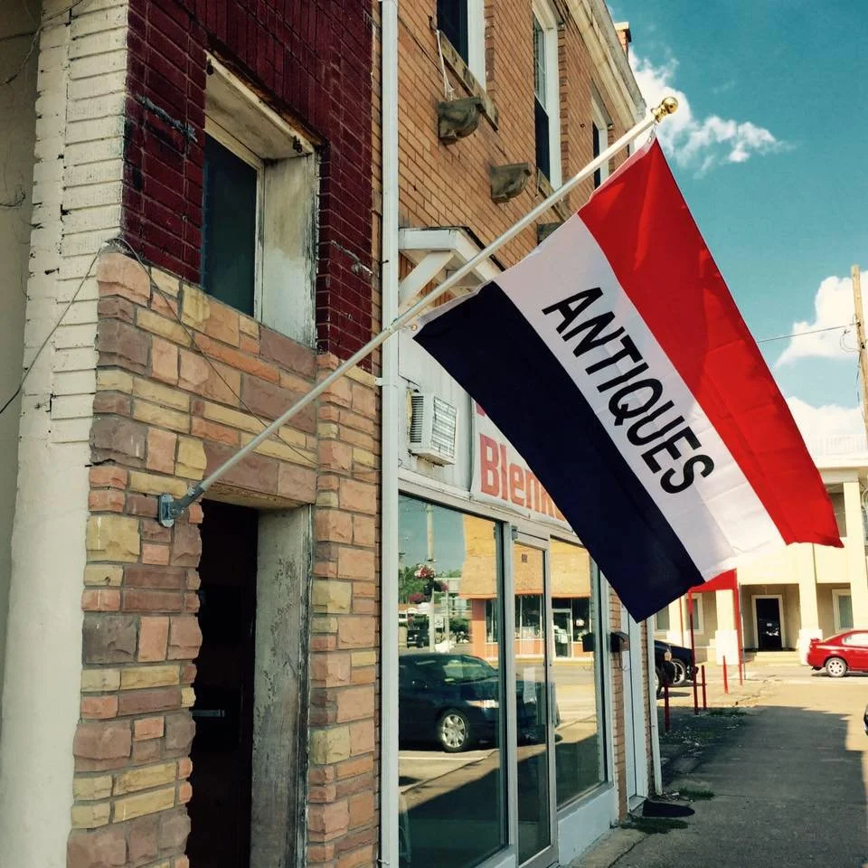 A red, white, and blue flag with the word "ANTIQUE" on it is mounted on the exterior of a brick building. The building has a large glass window and appears to be located on a street with parked cars and other buildings.
