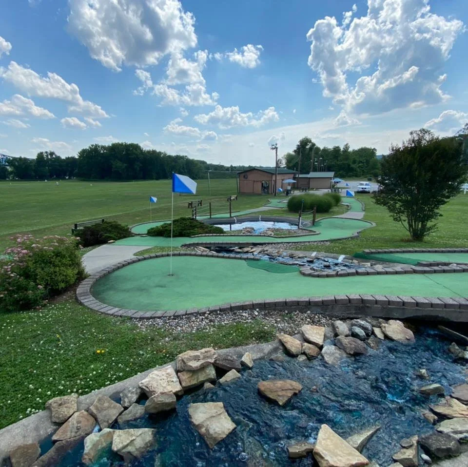 Mini golf course with strategically placed flags, small water features, and a bridge surrounded by green grass, bushes, and trees under a partly cloudy sky.