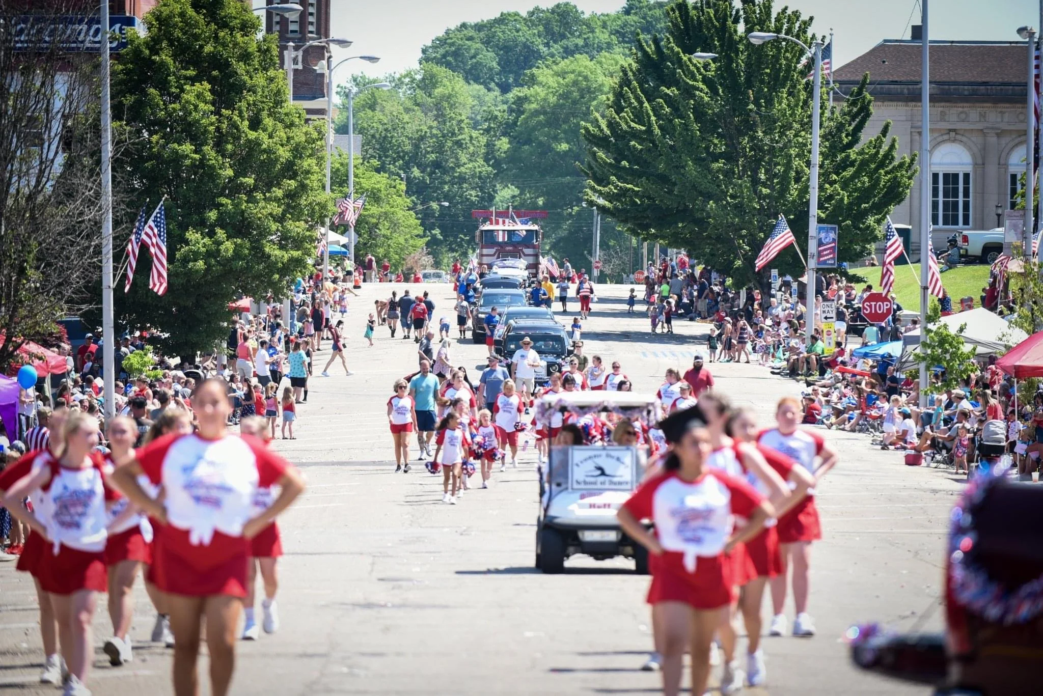 A parade on a sunny day with people lining the streets, many holding American flags. Participants, some dressed in red, white, and blue, walk along the street with a golf cart and a fire truck visible in the background. The street is decorated with American flags and festive banners, with large green trees lining the sides.