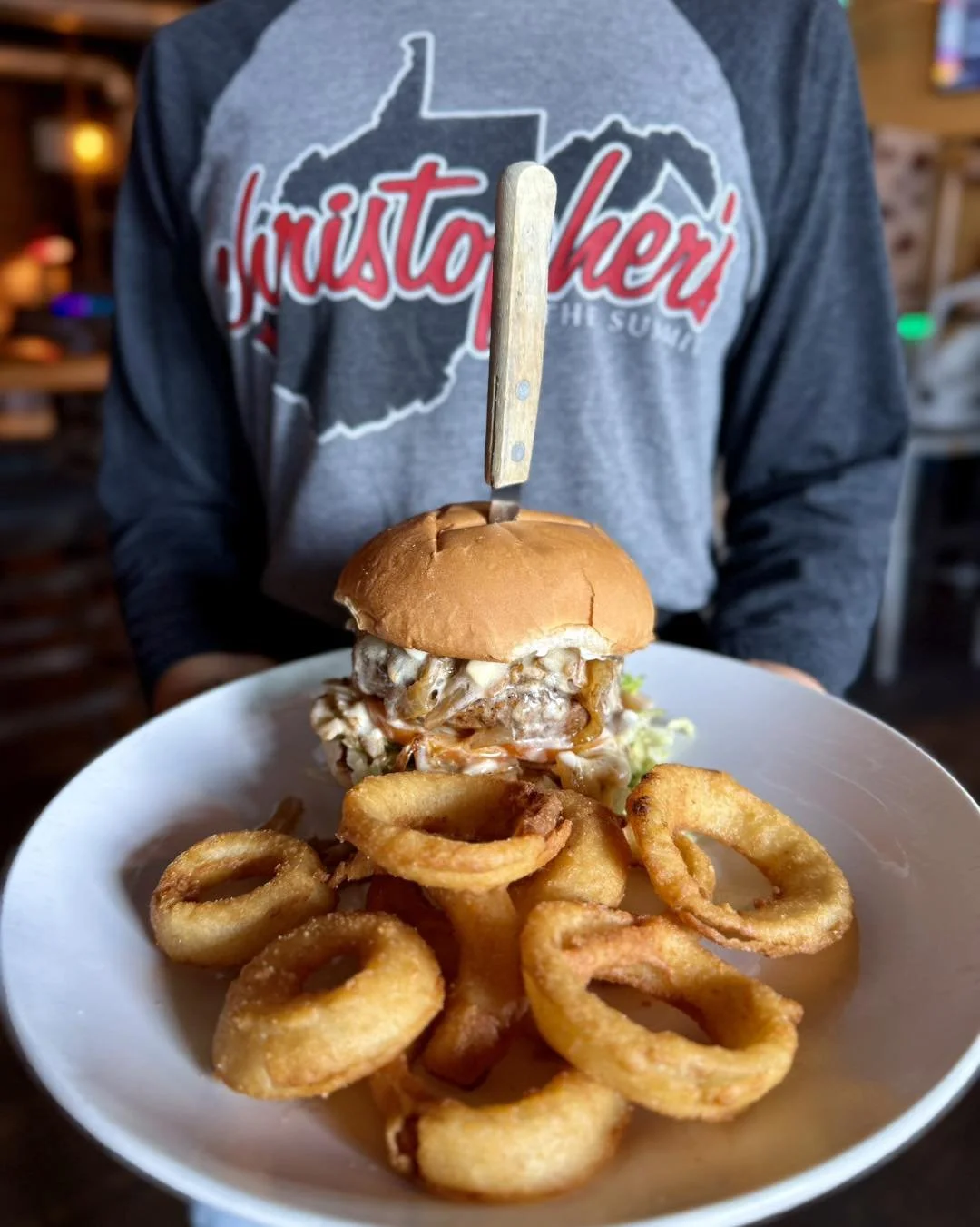A person holds a white plate with a burger topped with a knife, along with fried onion rings in the foreground. The person is wearing a hoodie with a West Virginia outline and the word 'huntington' in red.