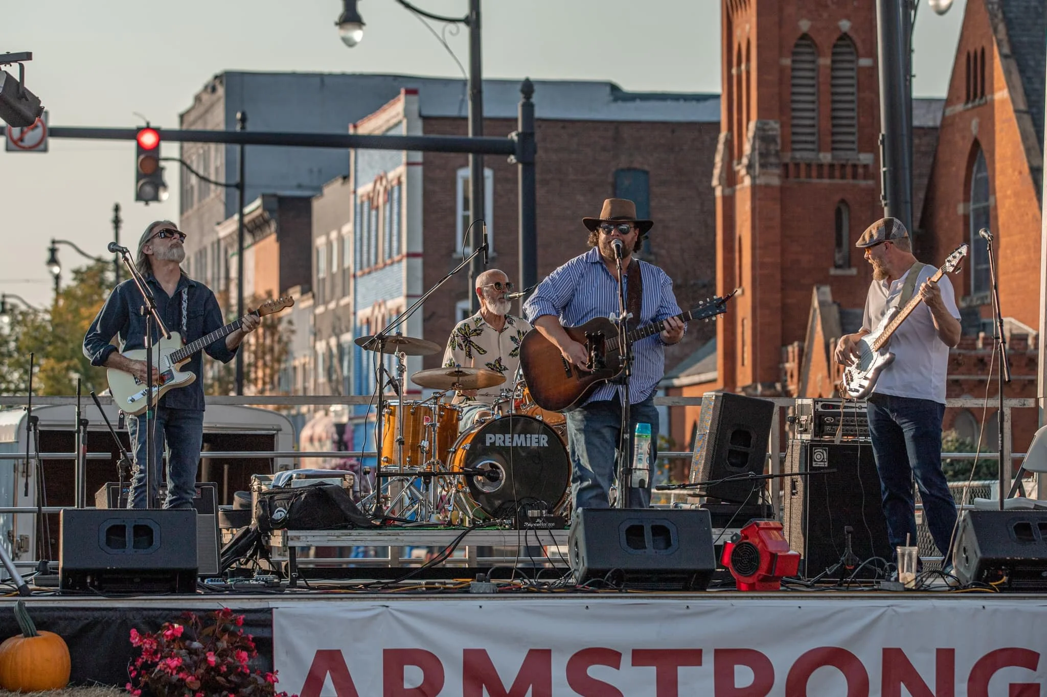 A live outdoor band performs on a stage with four members playing guitars and drums, set against a backdrop of historic brick buildings and a church, during daylight hours.