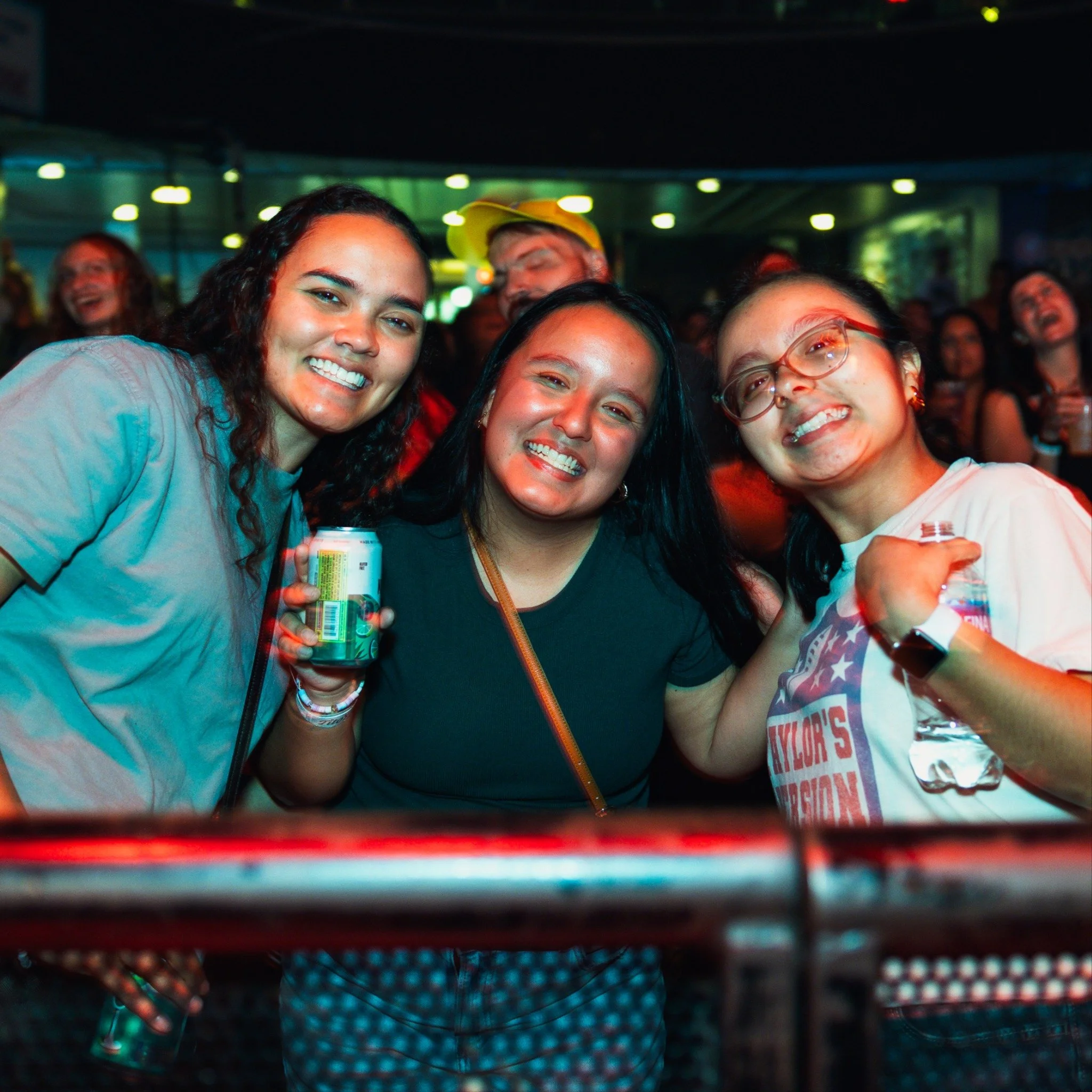 Three women smiling at a concert or event, one holding a beverage, with a crowd in the background.