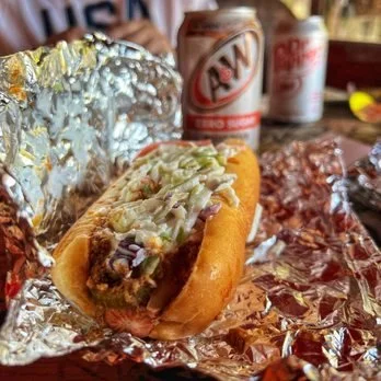 Hot dog topped with shredded lettuce and mustard, wrapped in foil, with cans of A&W root beer in the background.