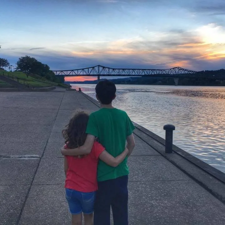 Two children, a girl and a boy, standing by a river at sunset with their arms around each other, looking towards a bridge in the distance.
