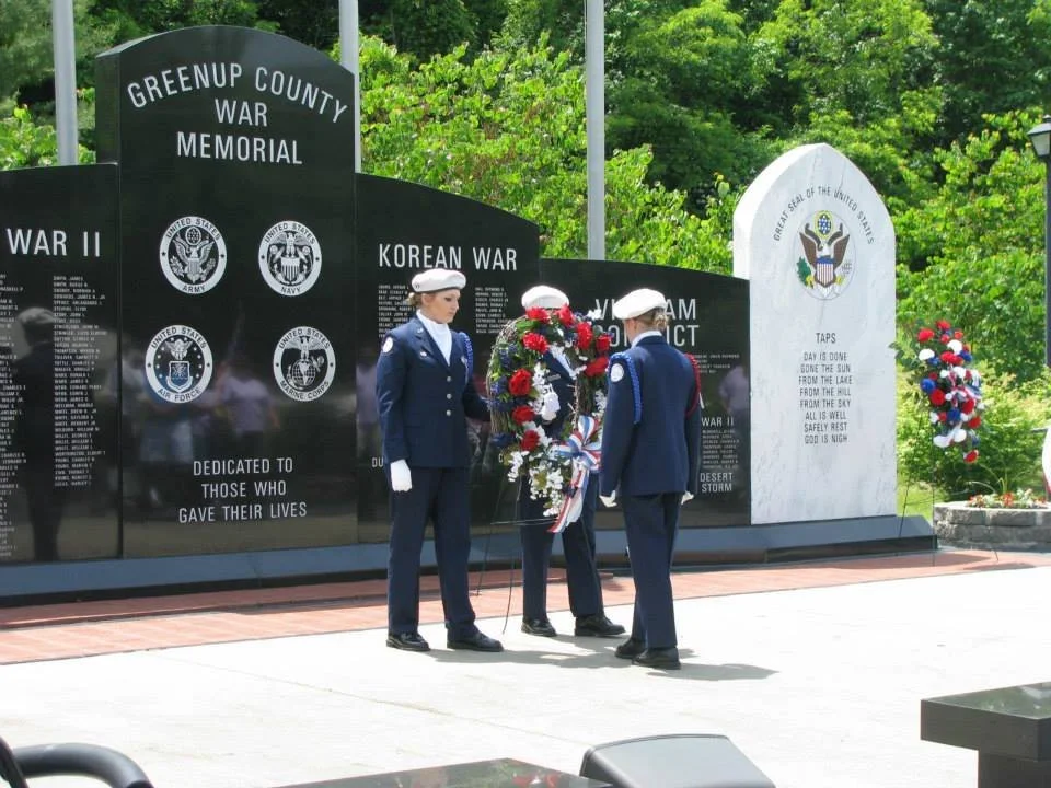 U.S. military personnel paying respects at the Greenup County War Memorial, with wreaths and a memorial wall in the background.