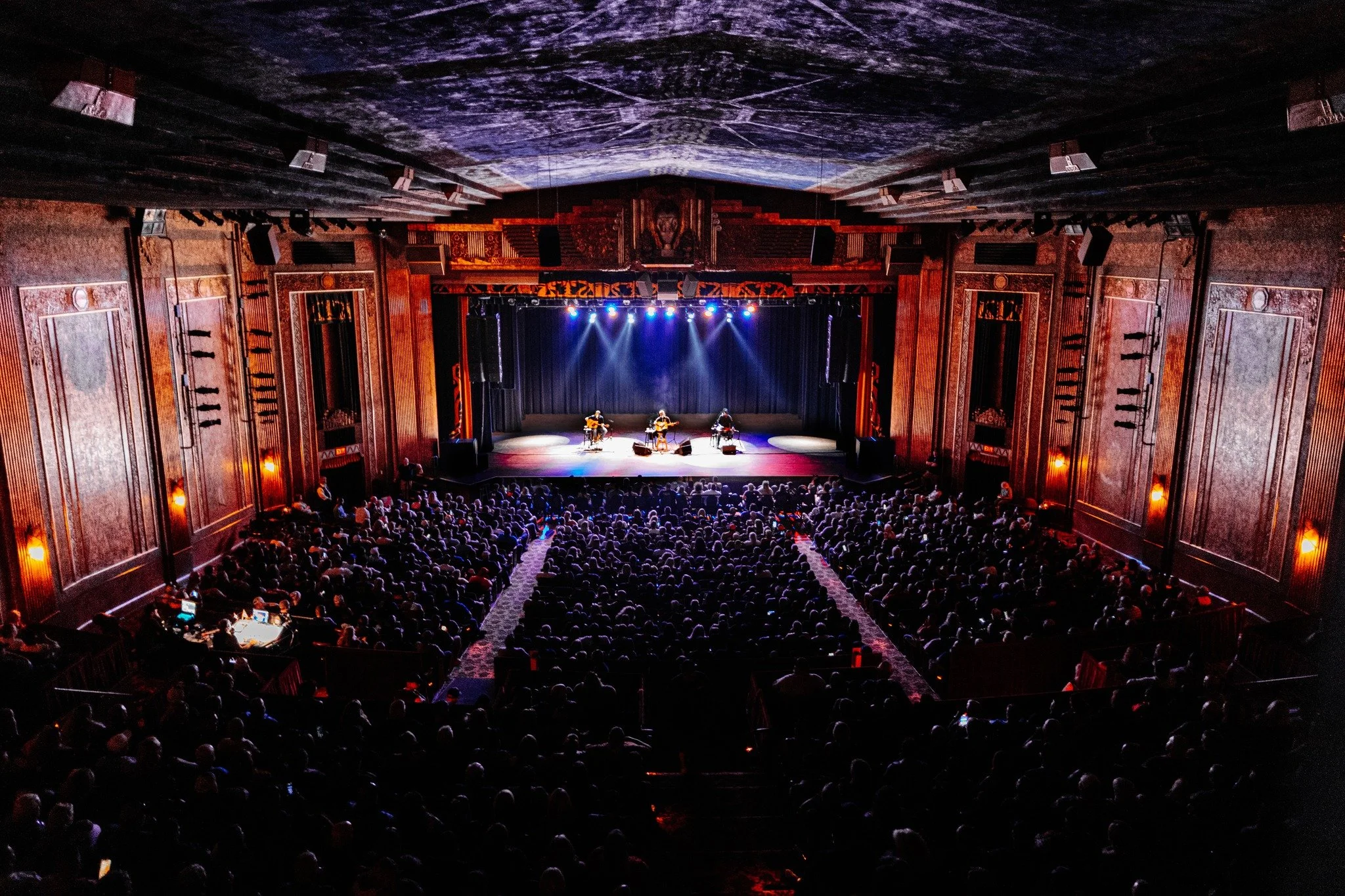 A large theater or concert hall filled with a seated audience watching a live musical performance on stage. The stage has three musicians playing guitars and one performing with a microphone, illuminated by stage lights.