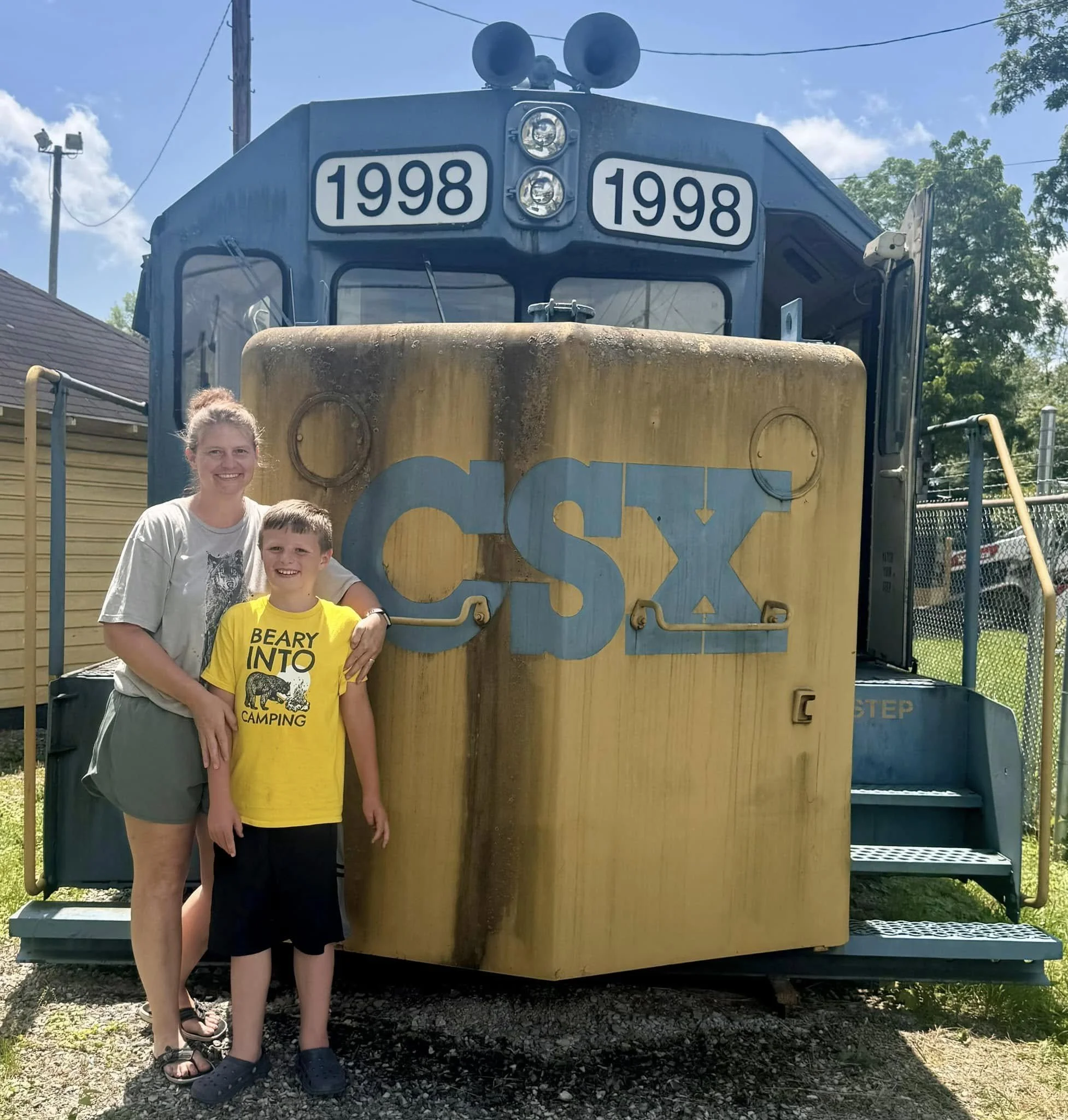 Two people, a woman and a boy, standing in front of a vintage train with the letters 'CSX' on the front and the year '1998' displayed above the train's windows. The woman is smiling and the boy is wearing a yellow T-shirt with a bear and camping graphic.