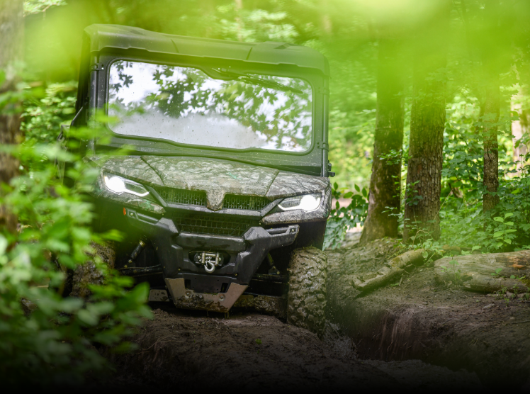 An off-road utility vehicle moving through a muddy trail in a dense, green forest.