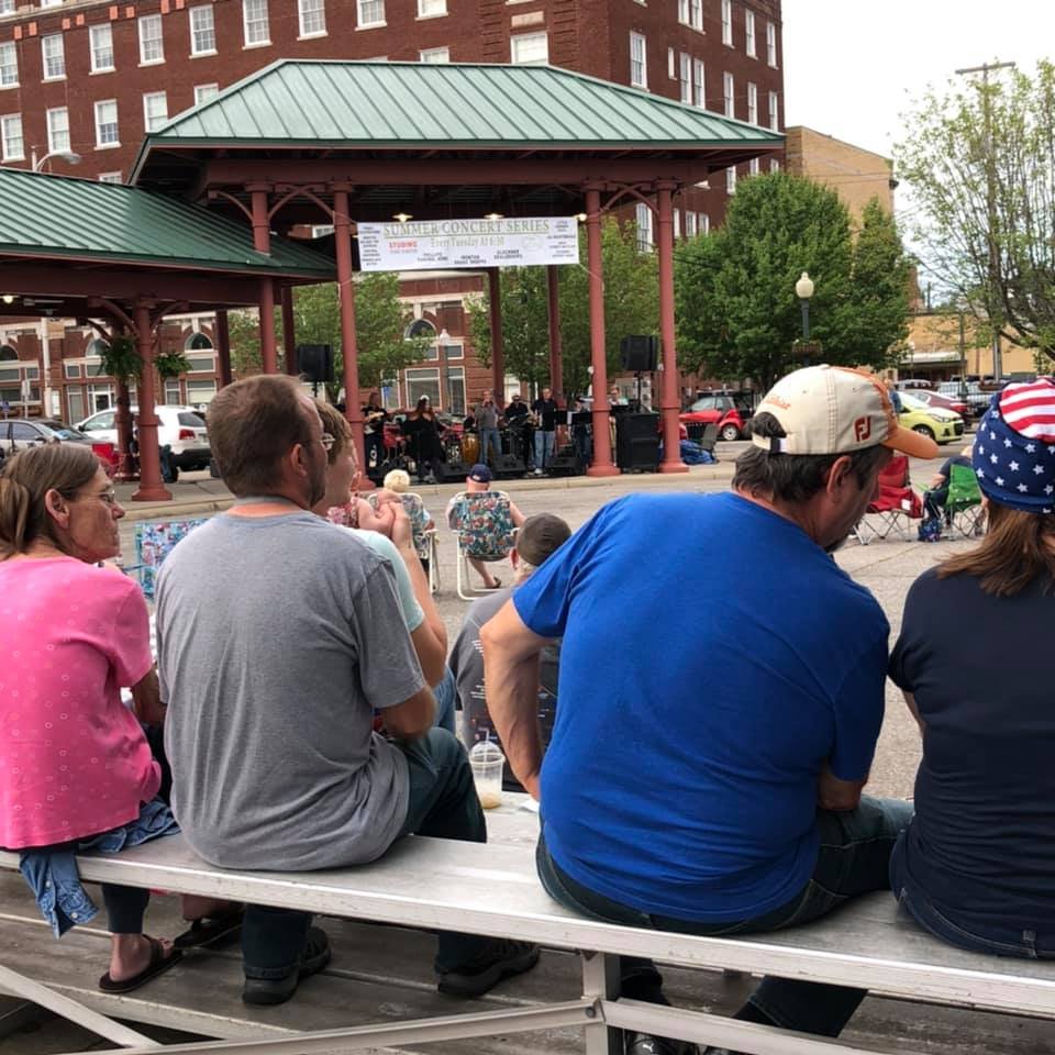People watching a band perform on an outdoor stage in a town square during a summer concert.