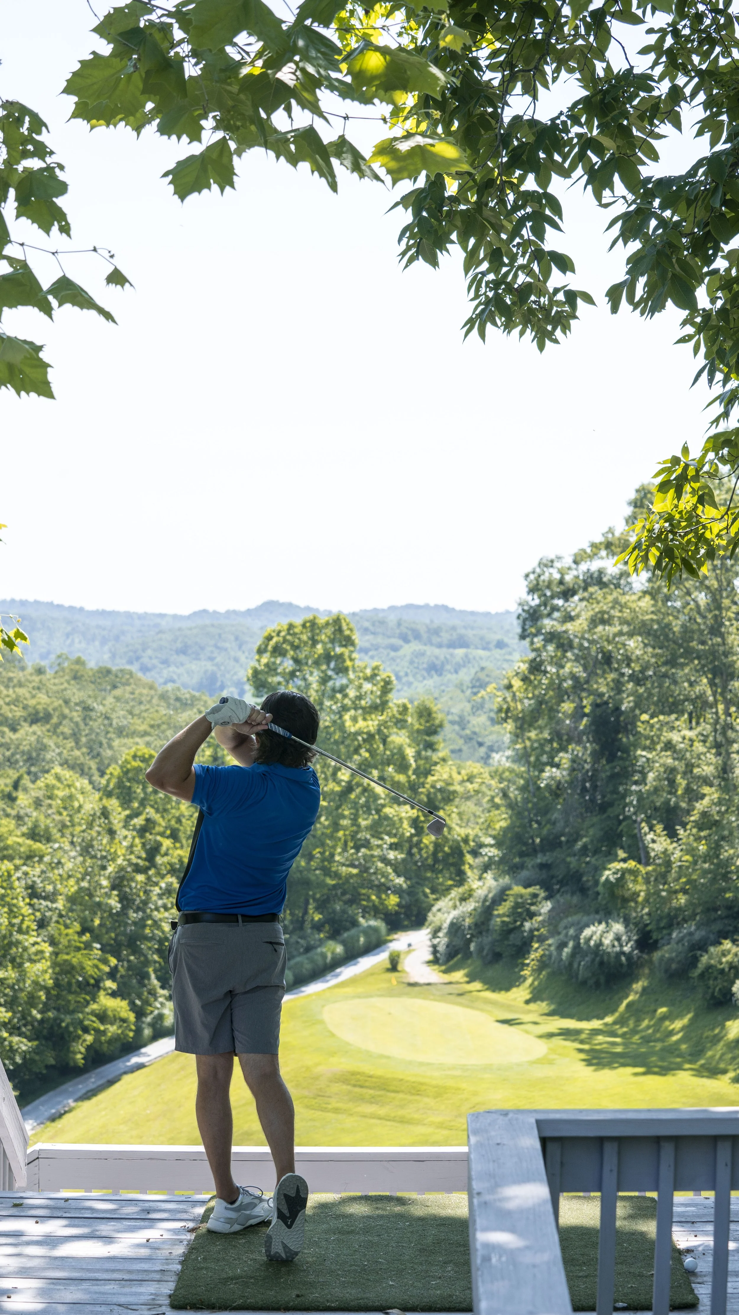 A man in a blue shirt and shorts is swinging a golf club on a golf tee box overlooking a lush, green golf course with trees and hills in the background.