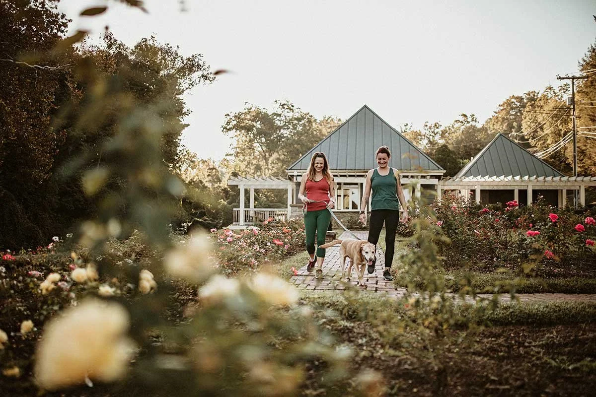 Two women walking a dog through a garden filled with blooming flowers, with a house in the background on a sunny day