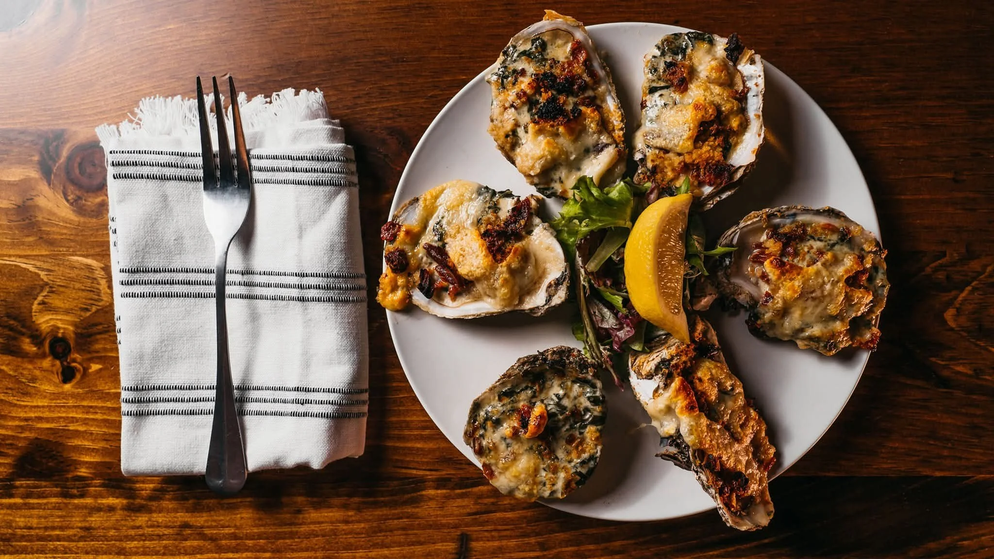 A white plate with six oysters topped with cheese, herbs, and bacon, garnished with a lemon wedge and mixed greens, placed on a wooden table. To the left, a fork is resting on a neatly folded white napkin with black stripes.