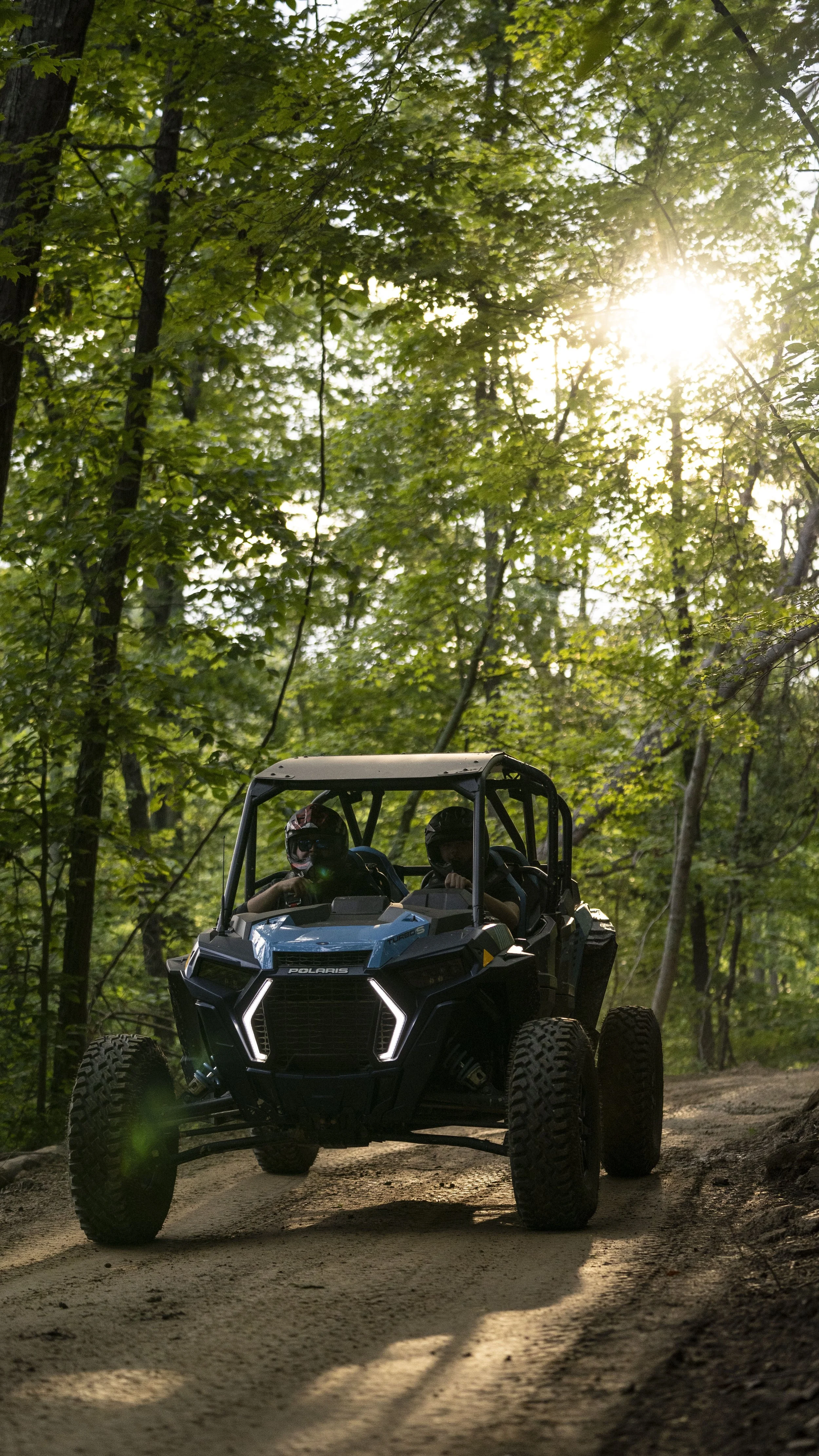 A Polaris off-road vehicle with two passengers wearing helmets driving on a dirt trail in a dense forest with sunlight filtering through the trees.