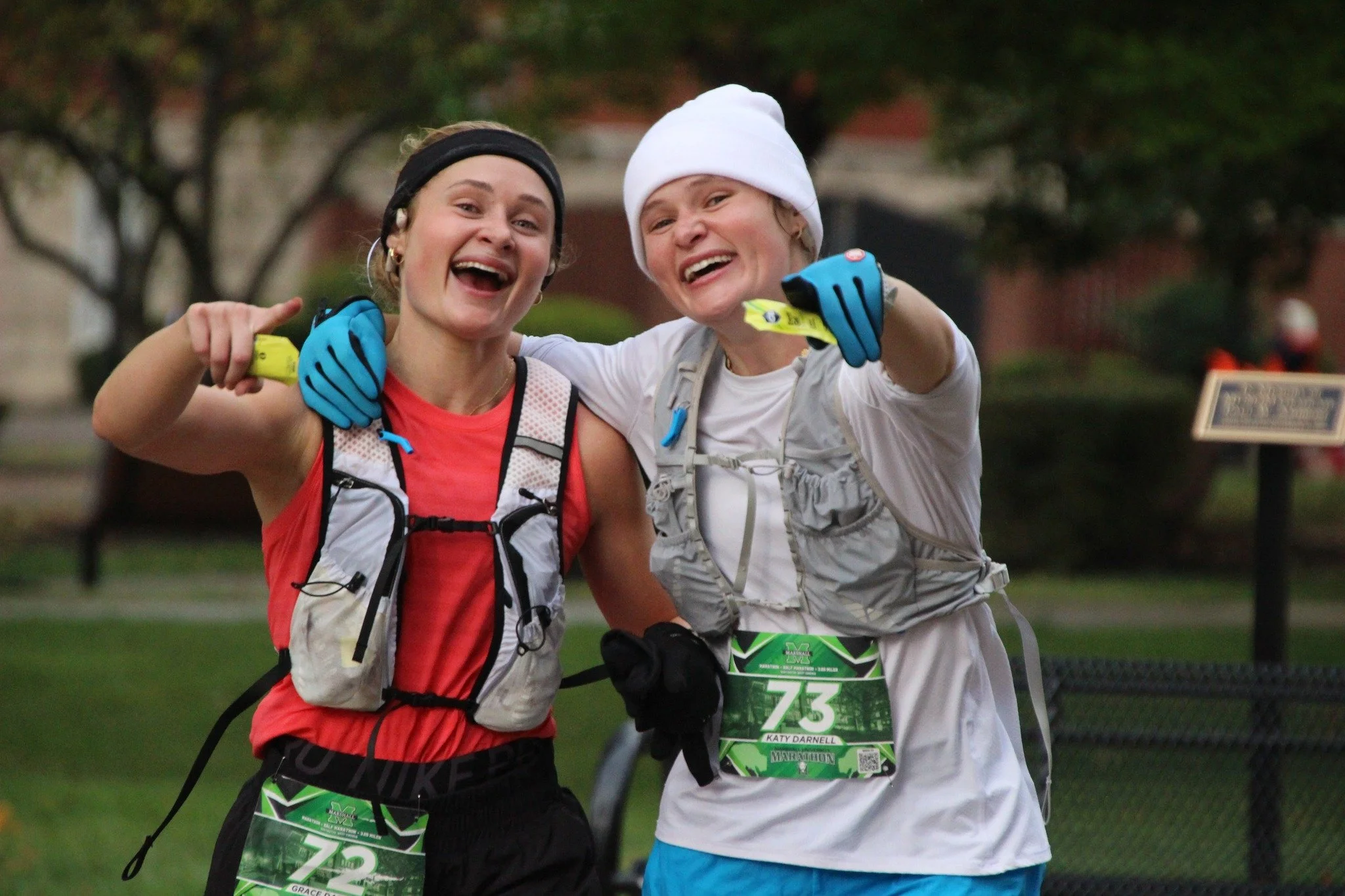 Two smiling women in athletic gear celebrate after a race, with race bibs and hydration packs, pointing towards the camera.