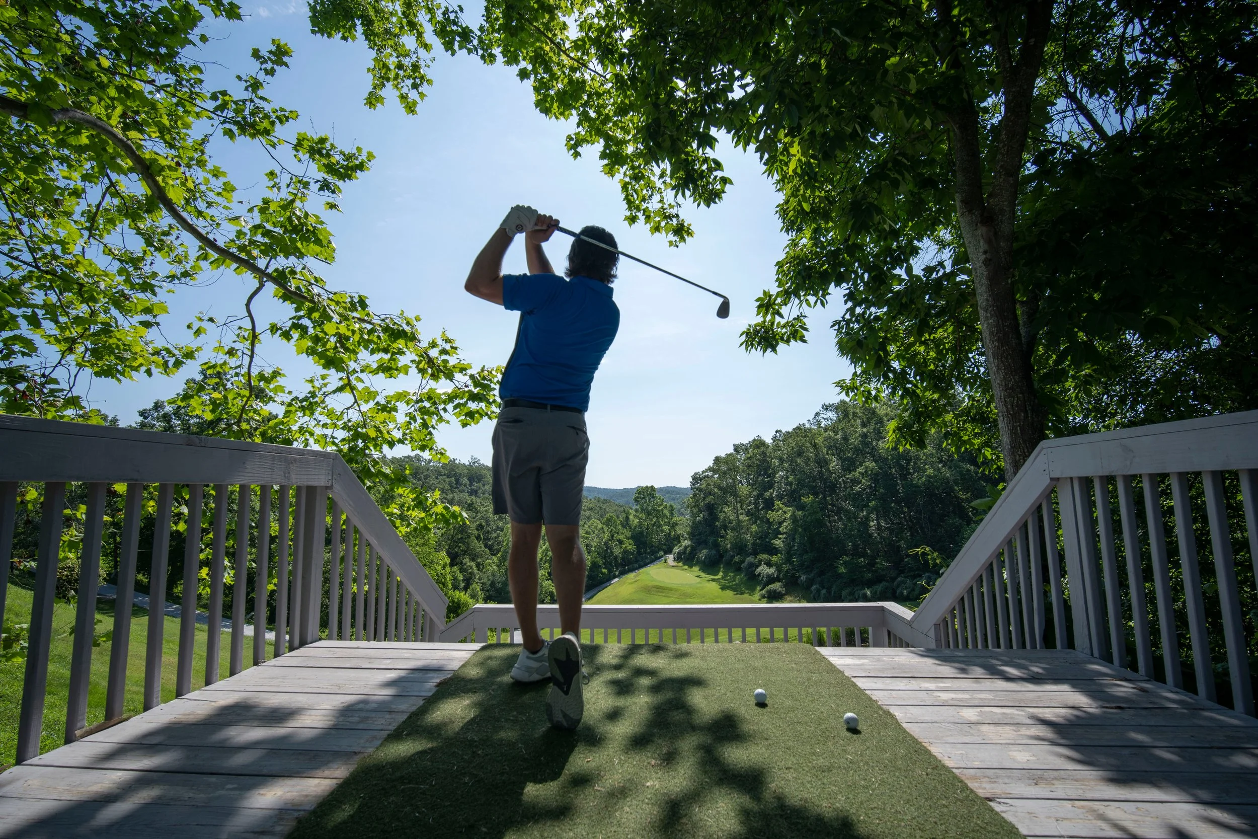 A man in a blue shirt and grey shorts playing golf on a wooden platform overlooking a lush green golf course surrounded by trees on a sunny day.