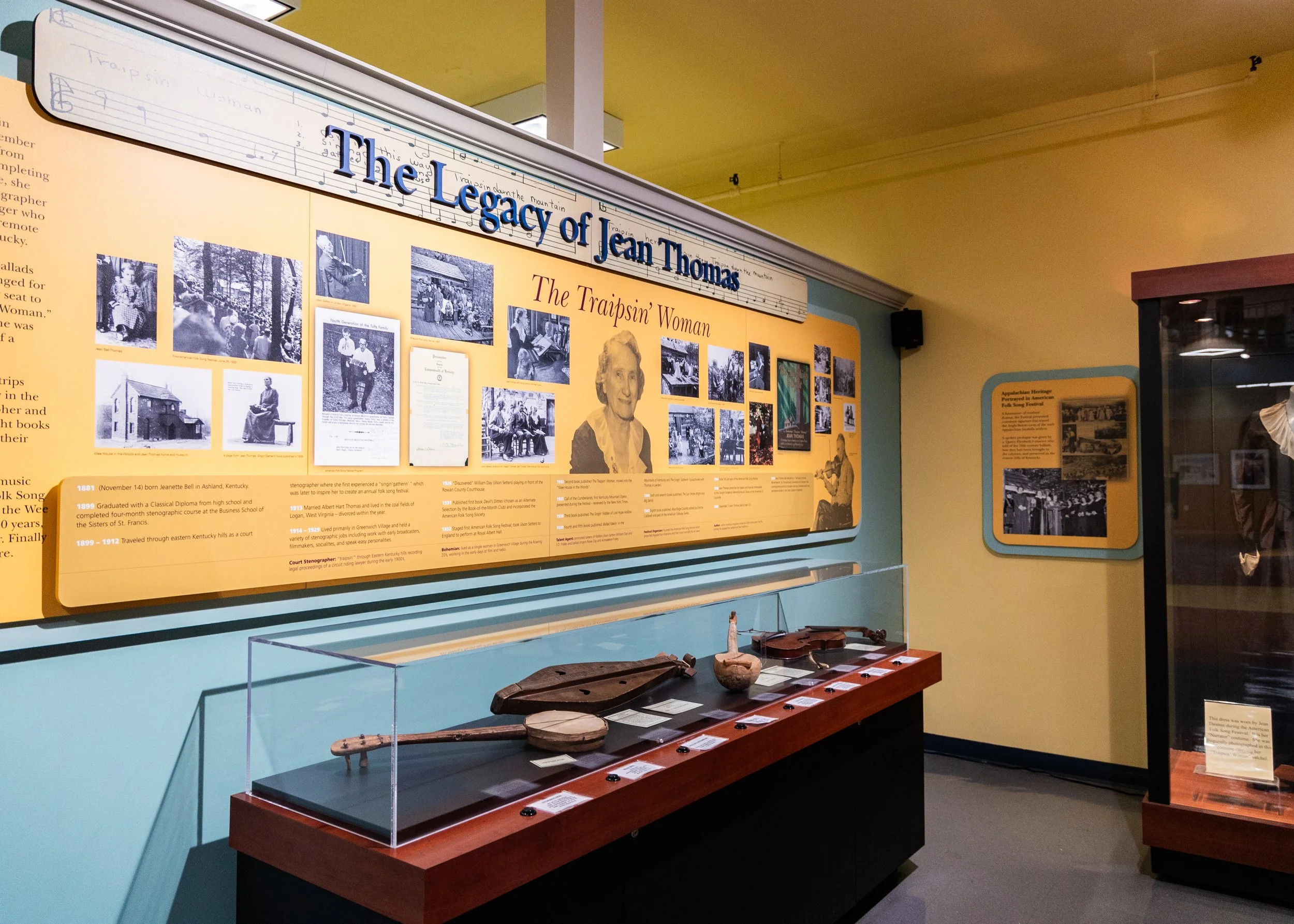 Exhibit display at a museum dedicated to Joan Thomas, titled "The Legacy of Joan Thomas." The display includes a large yellow informational panel with black and white photographs, and artifacts such as wooden objects encased in glass beneath it.