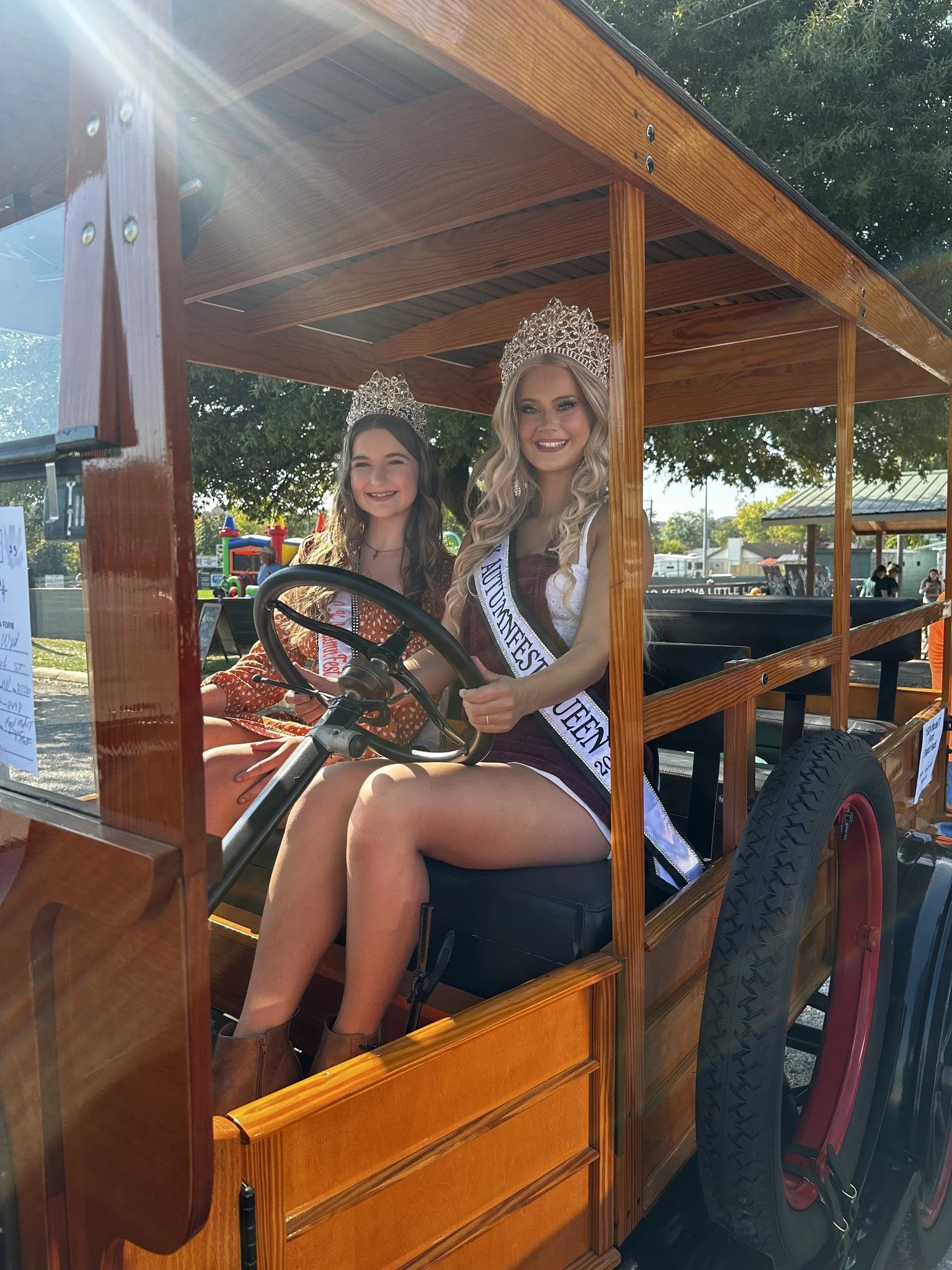 Two young women wearing pageant crowns and sashes, sitting in a wooden parade float with a steering wheel, at an outdoor event on a sunny day. One is the queen, and the other is the princess, smiling at the camera.