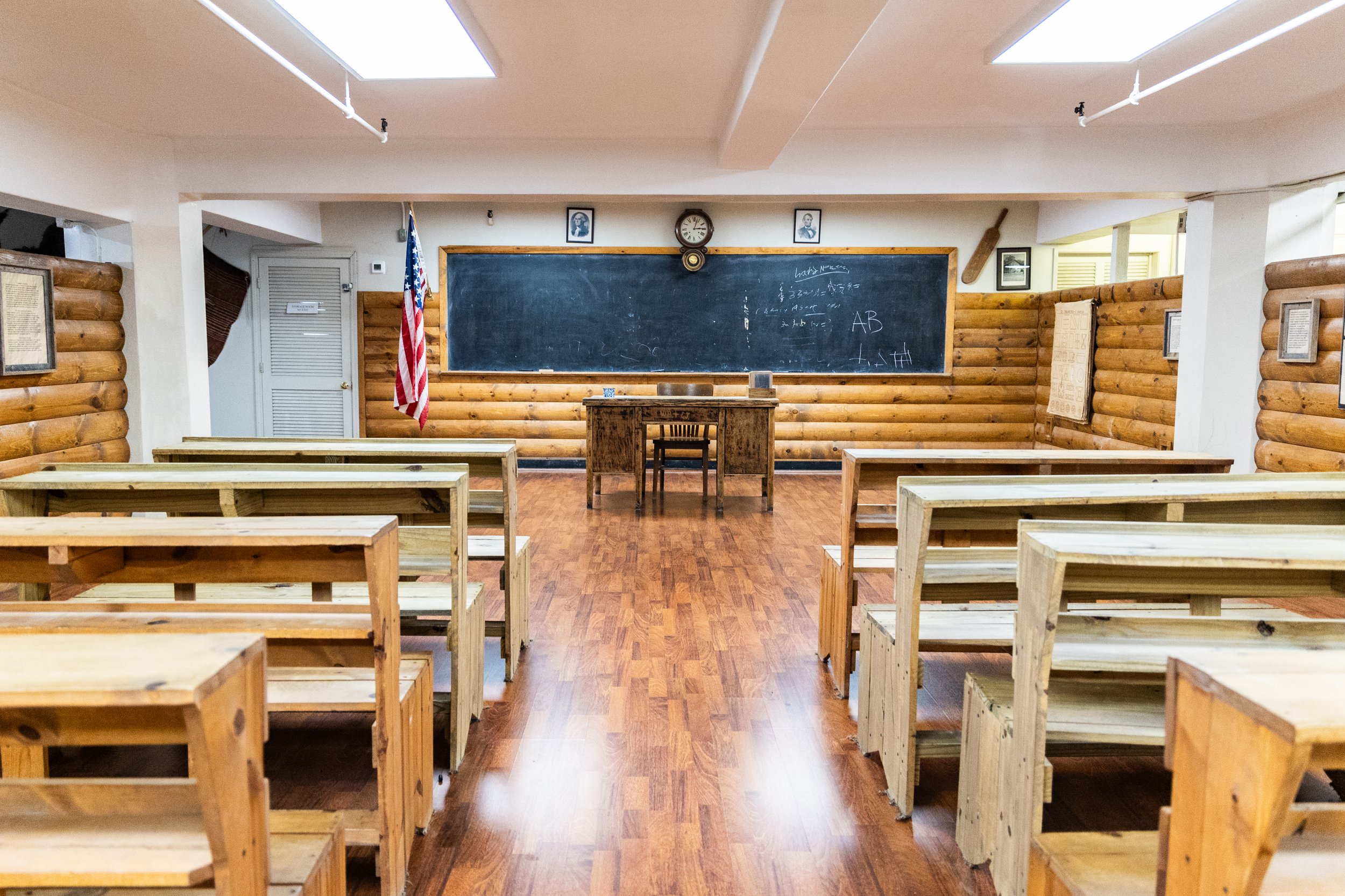 Empty classroom with wooden desks, a blackboard, an American flag, and framed pictures on the walls.