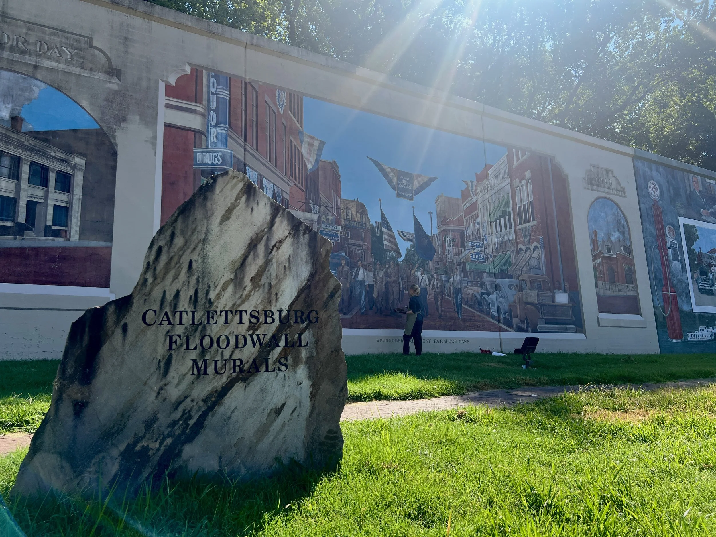 A large mural depicts a historic street scene with buildings, flags, and people, with a stone in the foreground engraved with "Cattletsburg Floodwall Murals." The mural is outdoors, with sunlight and green grass in front.