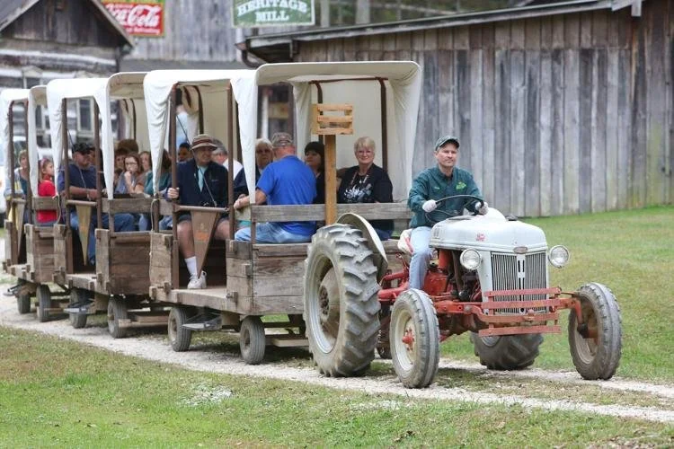 Group of people riding in a small open-air tractor-pulled train on a gravel path near a wooden barn.