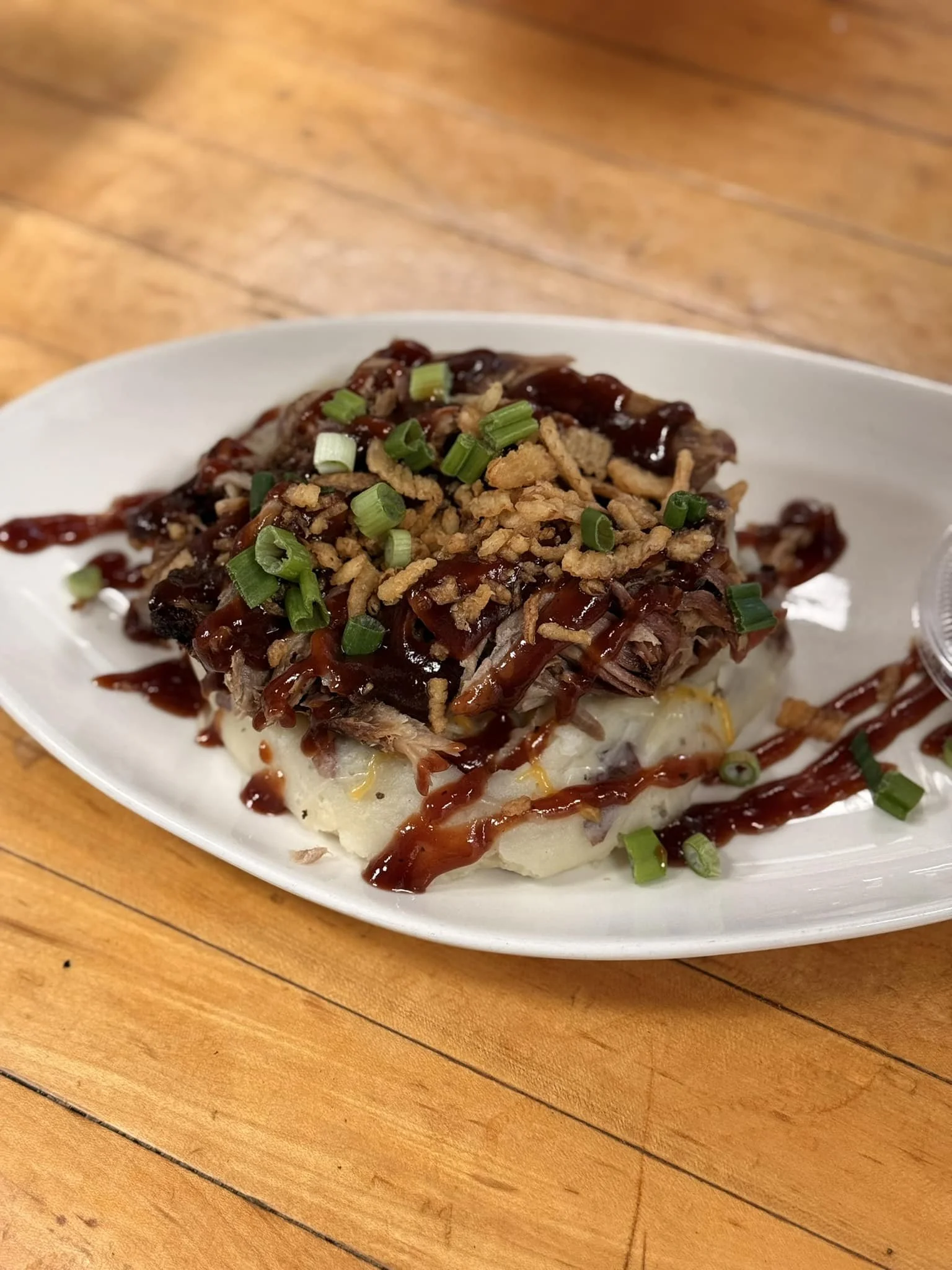 A plate of mashed potatoes topped with shredded meat, barbecue sauce, chopped green onions, and crispy shallots on a wooden table.