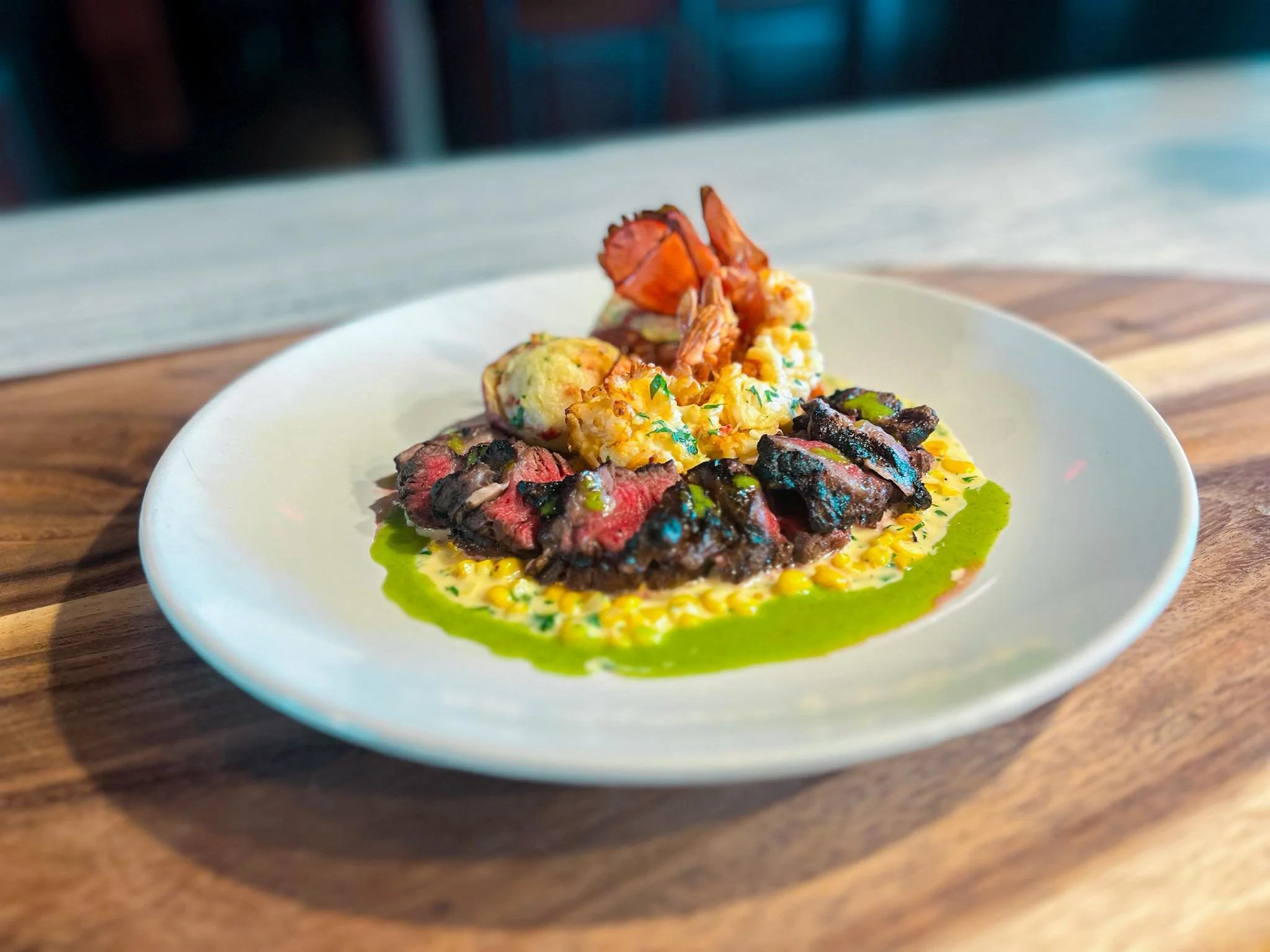 A white plate with sliced medium-rare steak, corn, and grilled shrimp, topped with green sauce, all on a wooden table.