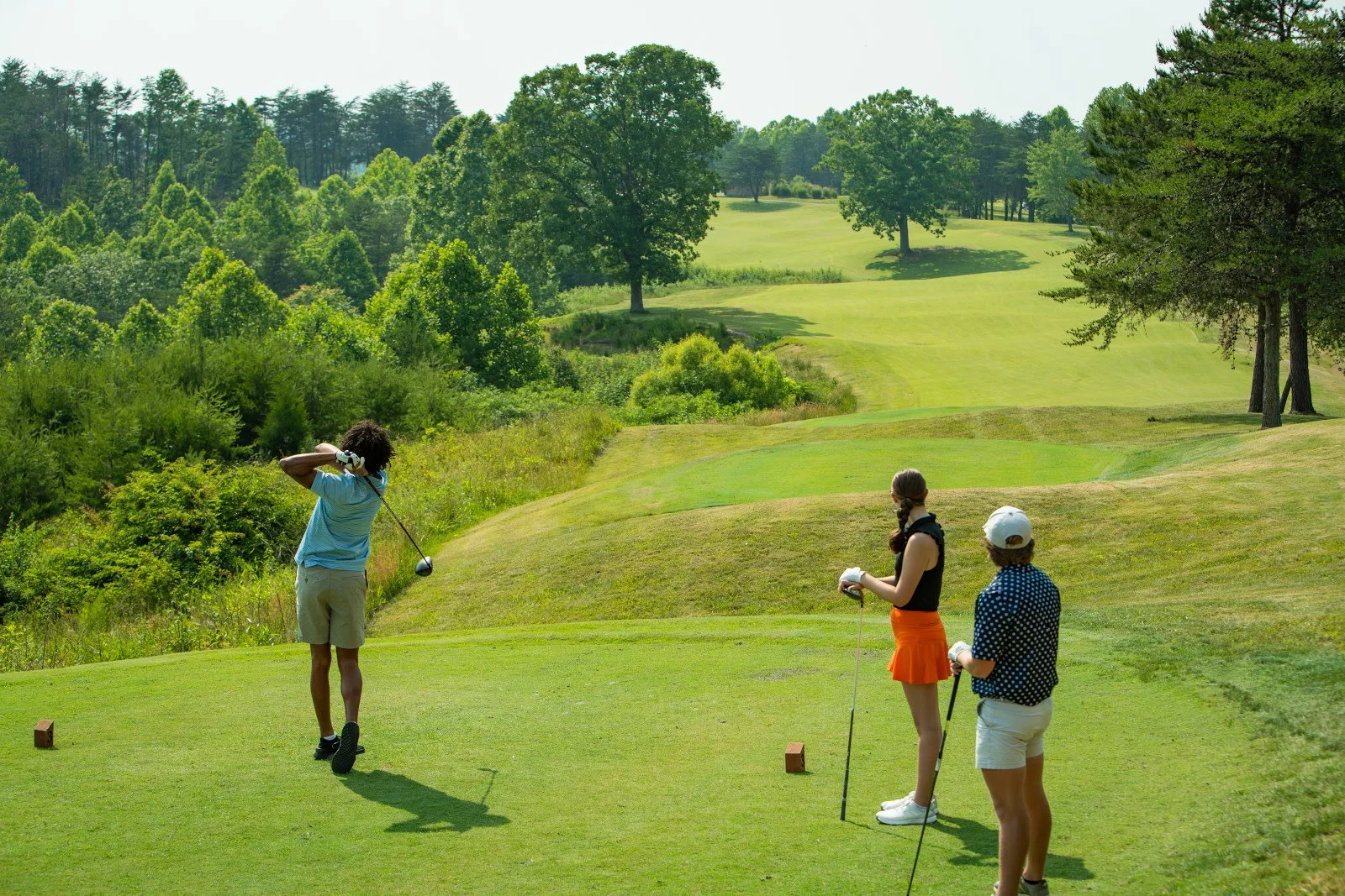 Three people preparing to hit golf balls on a lush golf course surrounded by trees and rolling hills.