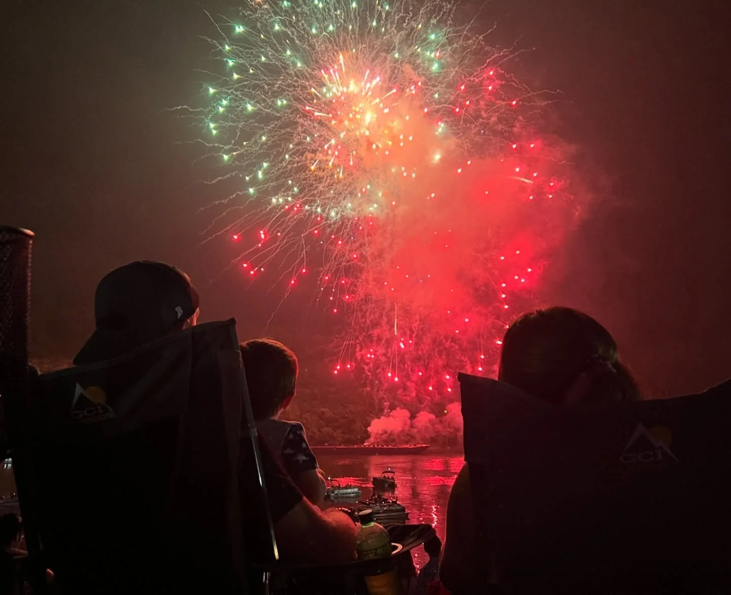 People sitting in chairs overlooking a body of water at night, watching fireworks display in the sky with bright colors and sparks.