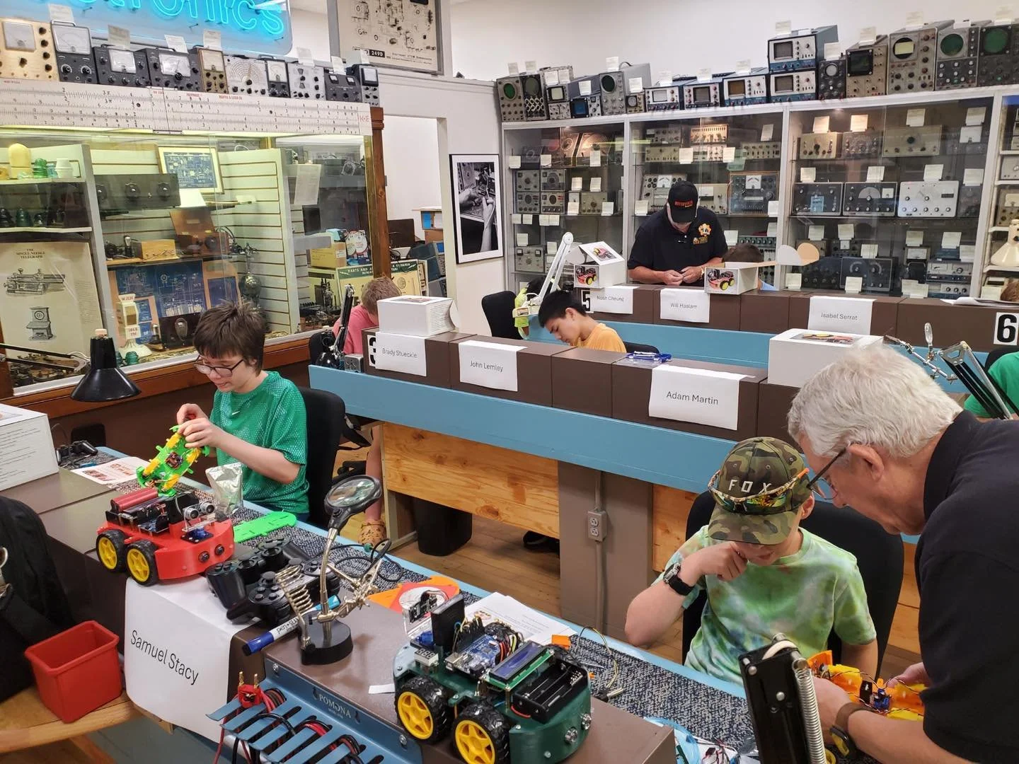 Children and adults participate in a robotics workshop at a museum, with exhibits of vintage electronic equipment and instruments displayed on shelves in the background.