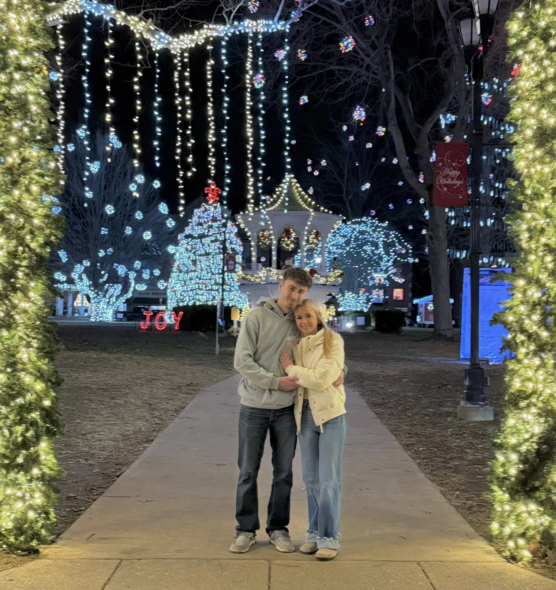 A young couple stands close together on a decorated holiday pathway at night, surrounded by Christmas lights and illuminated trees, with a festive gazebo in the background and the word 'JOY' lit up in red.