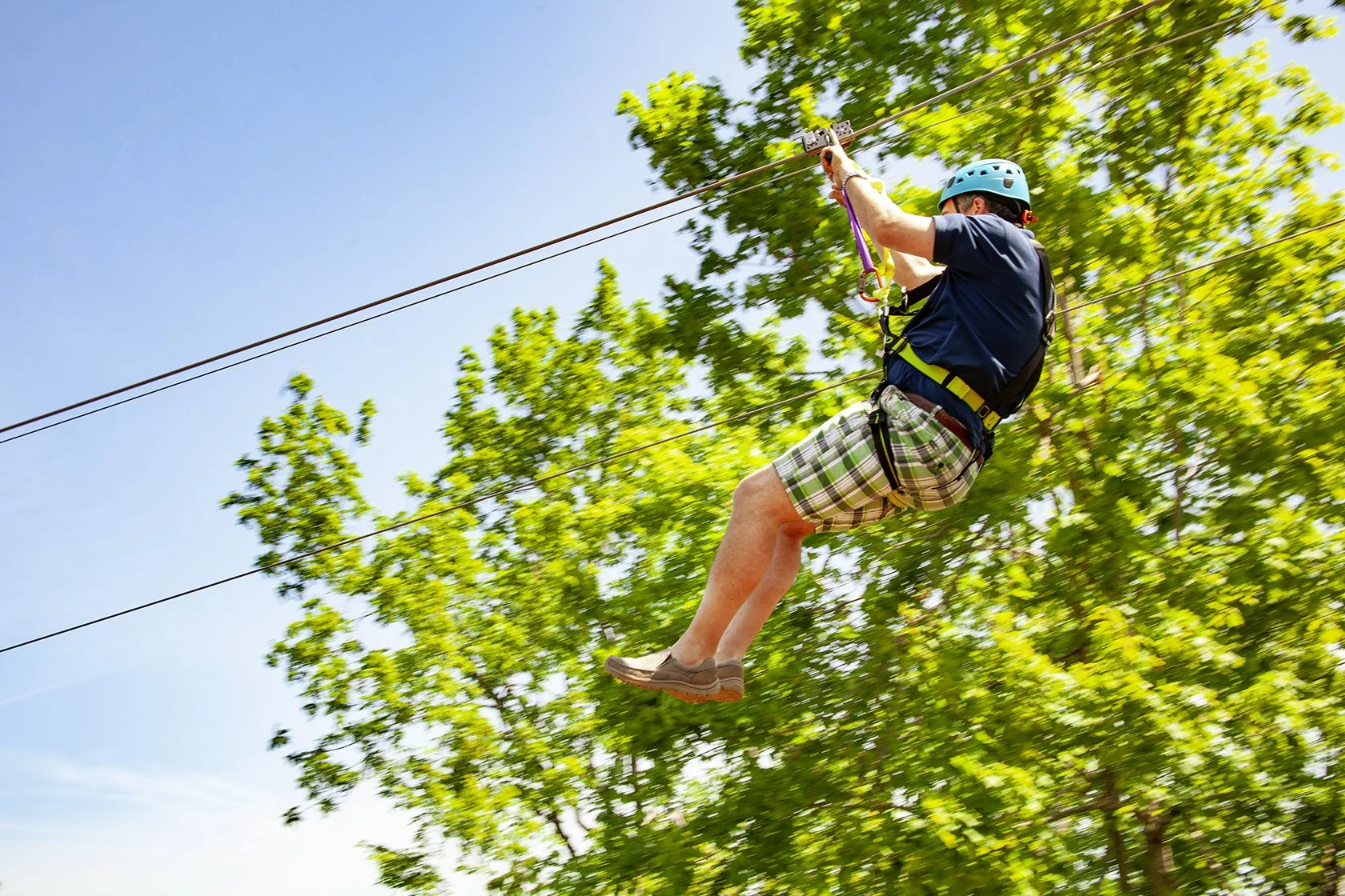 Man wearing a helmet and harness zip-lining through green trees on a sunny day.