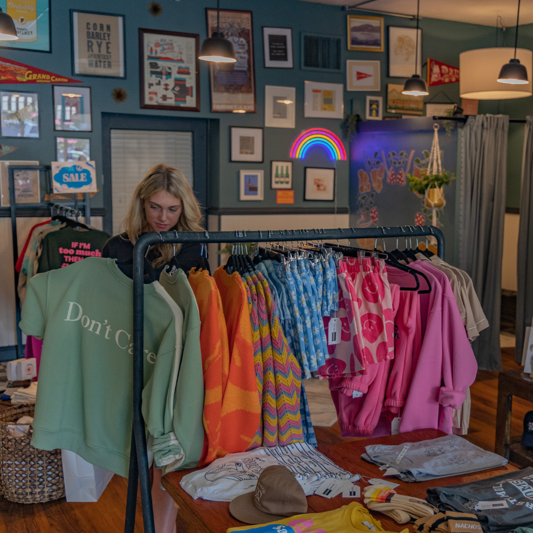 A clothing store with a woman looking at colorful sweatshirts on a rack. The store has framed art and neon signs on the wall, including a rainbow and Superman symbols. Various clothing items and hats are displayed on tables and racks.