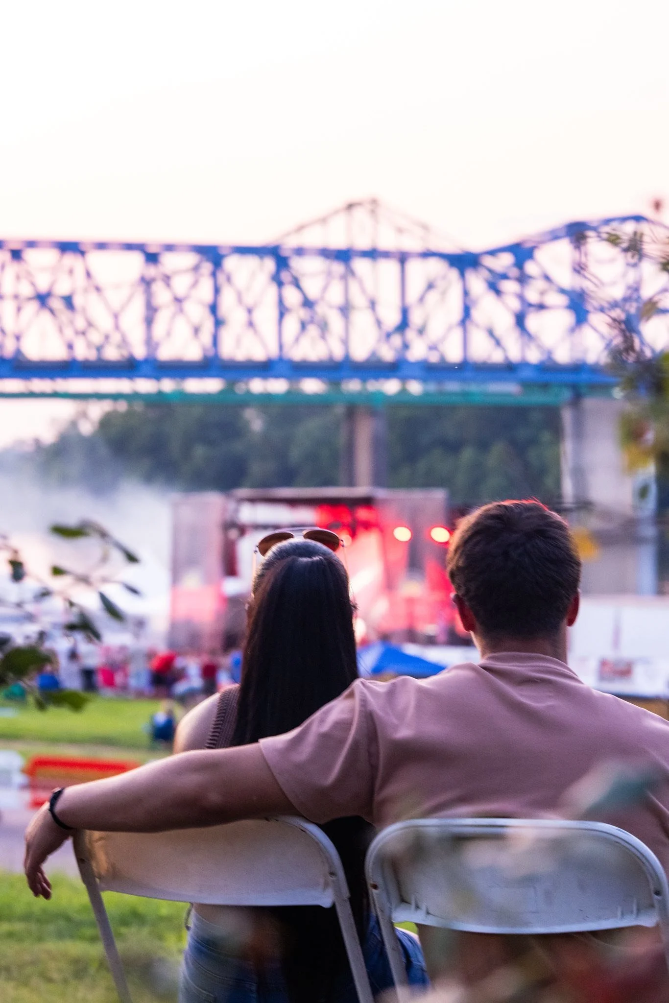 A man and a woman sitting outdoors watching a concert stage at dusk.