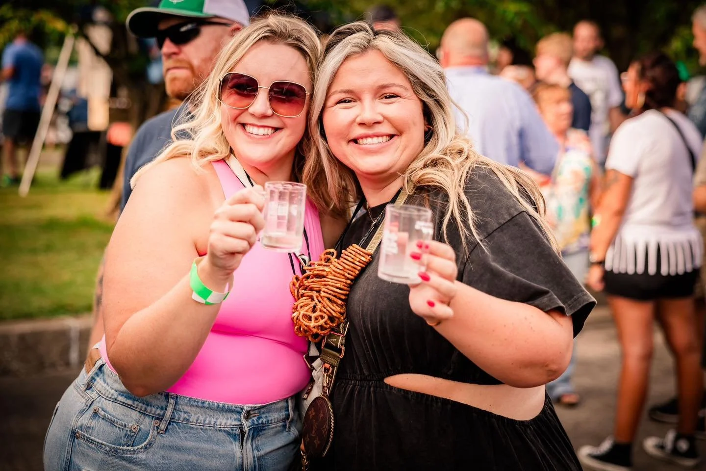 Two smiling women holding shot glasses at an outdoor event with a crowd in the background.