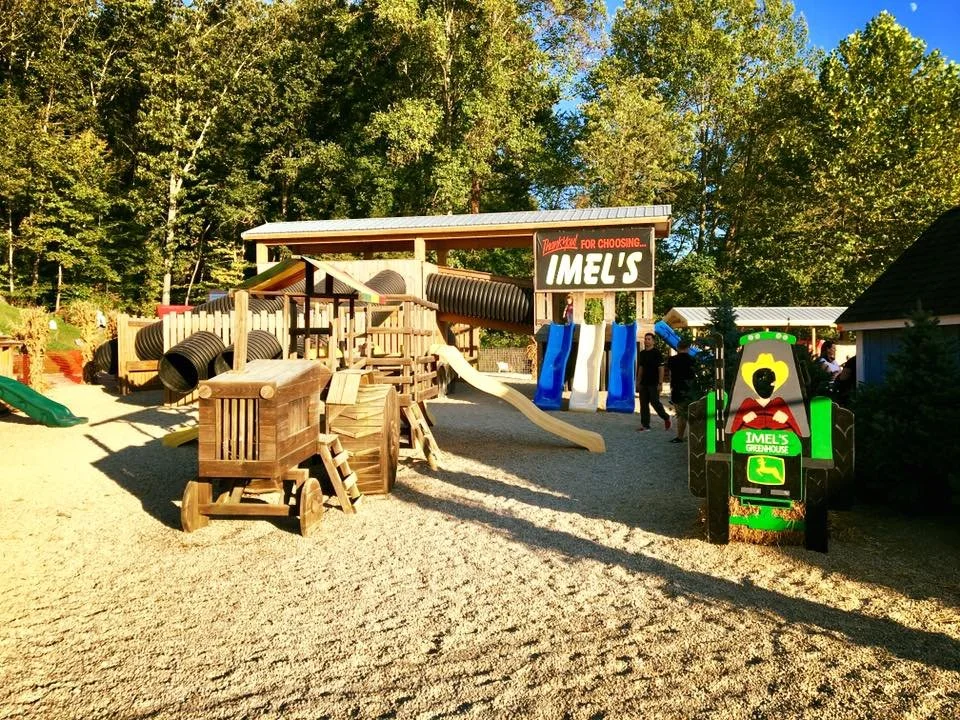 Children's outdoor playground at Imel's Greenhouse includes wooden structures, slides, tunnels, and a small playhouse, surrounded by trees.