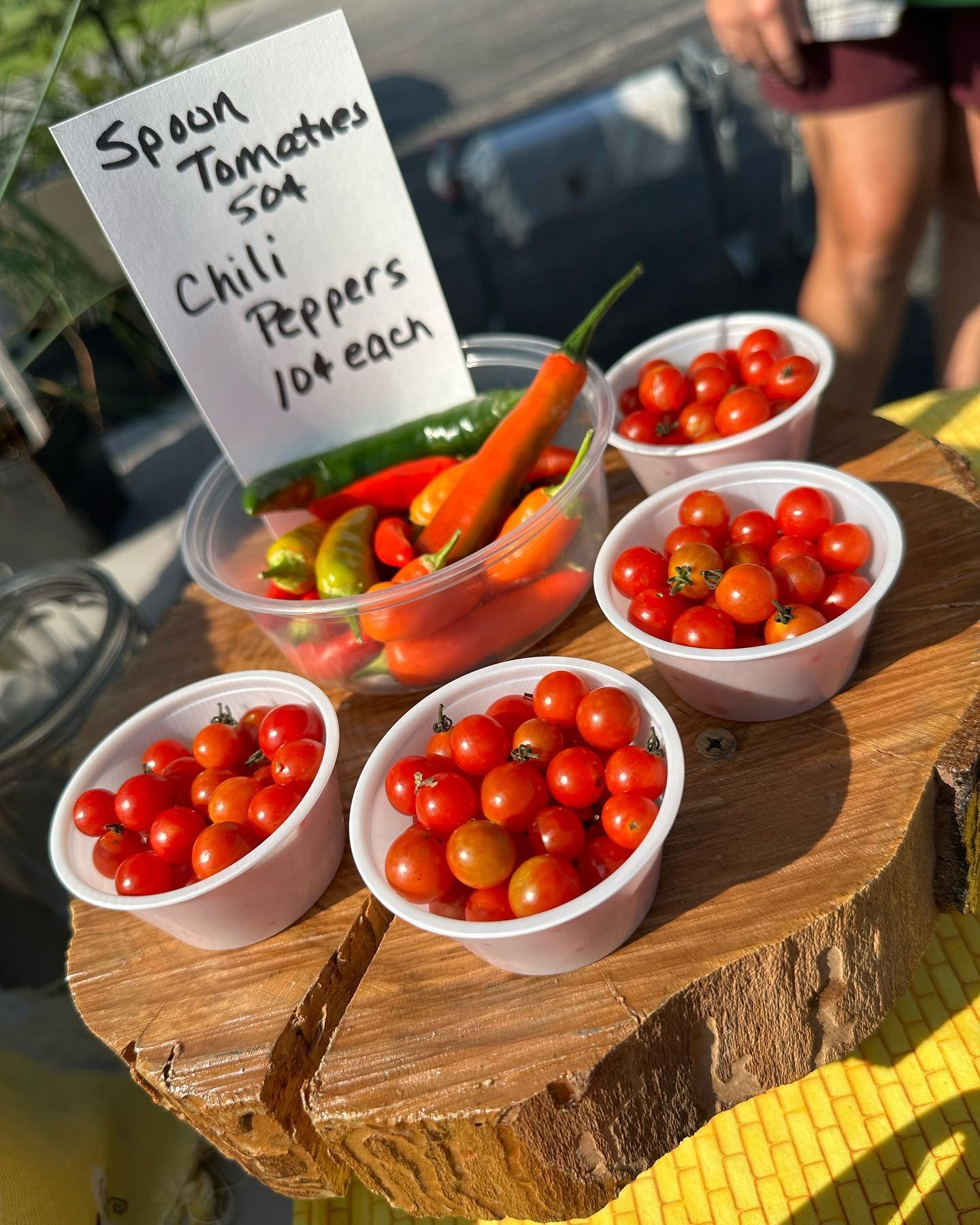 Cherry tomatoes in white cups, and hot peppers in a clear bowl with a handwritten sign listing other vegetables for sale.