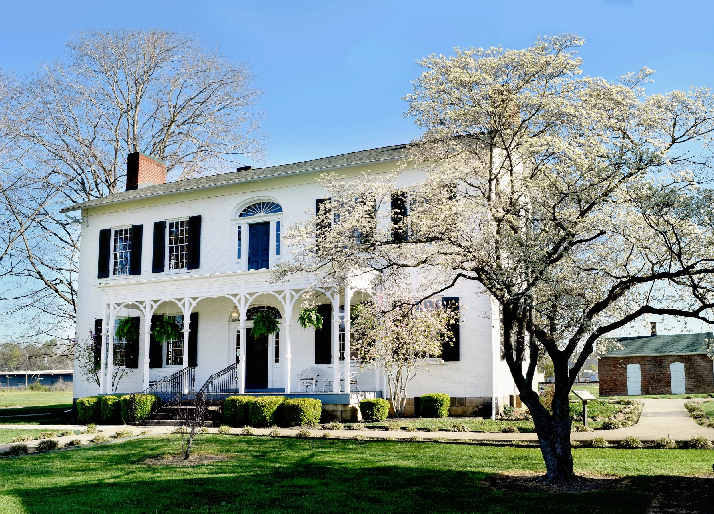 White two-story house with black shutters, a blue door, a covered porch with hanging plants, surrounded by greenery and a flowering tree in front, under a clear blue sky.