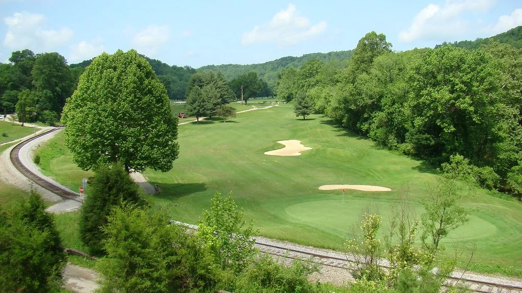 A golf course with green grass, sand bunkers, and trees, surrounded by hilly landscape and a railway track on the left side.