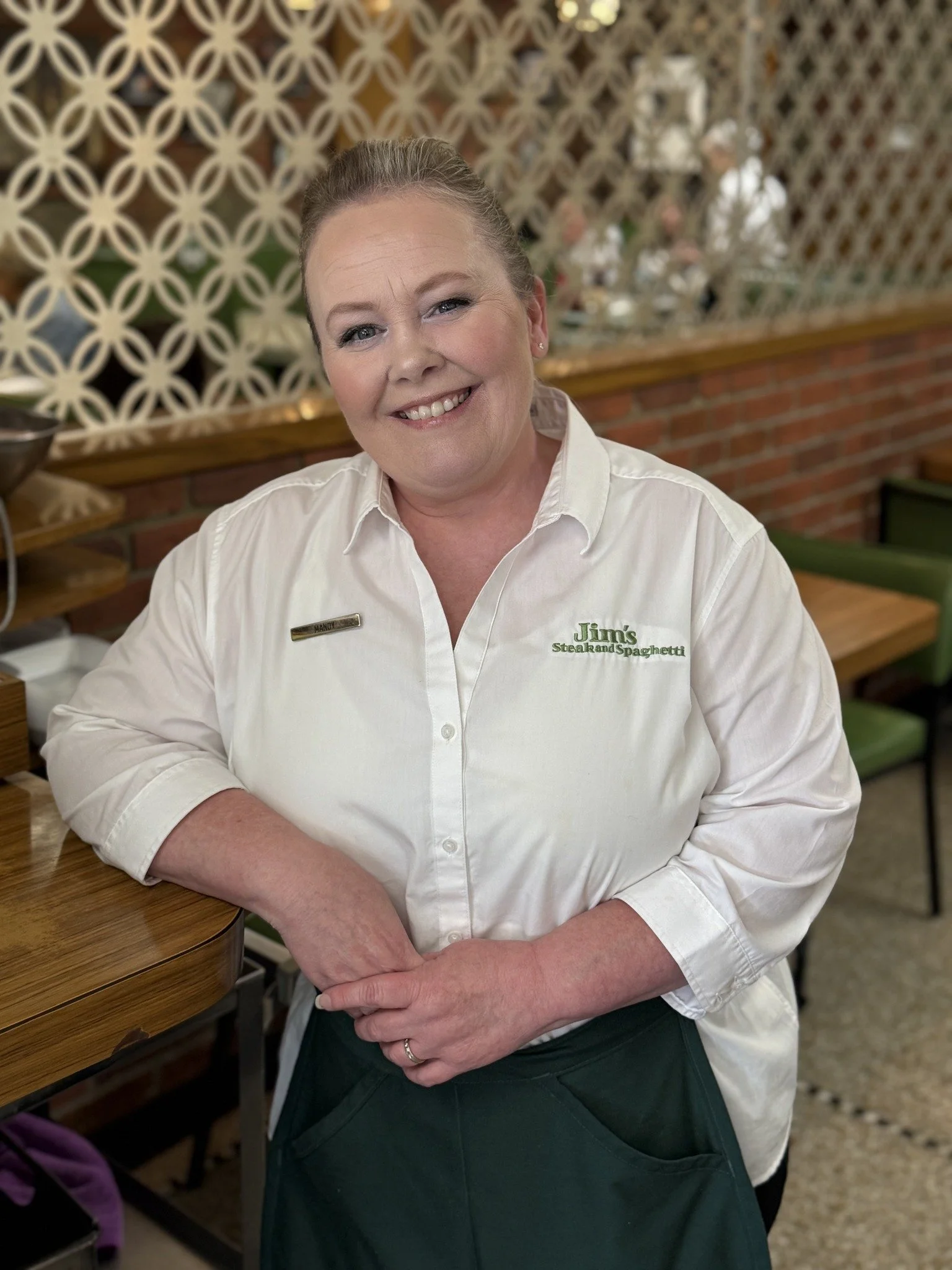 A smiling woman in a white shirt with a name tag and embroidered text reading 'Jim's Steak and Spaghetti' stands in a restaurant.