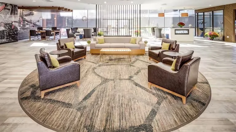 Modern hotel or lounge lobby with circular area rug, four leather armchairs with green pillows, a beige sofa with yellow pillows, and a white sofa at the back. Large windows let in natural light, and there are flowers and modern decor throughout.