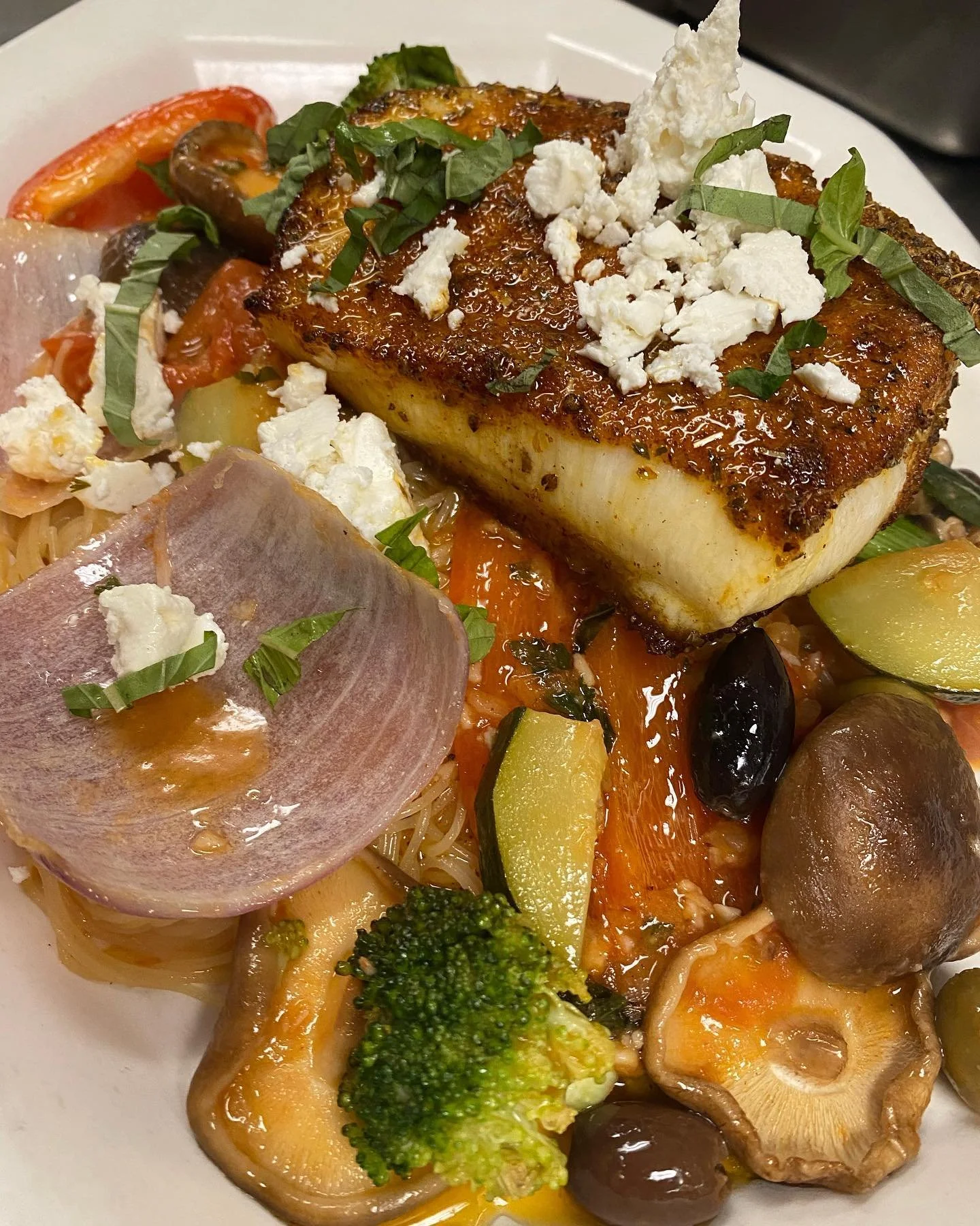 A plate of grilled fish fillet topped with chopped herbs and crumbled cheese, served with various cooked vegetables including broccoli, mushrooms, zucchini, tomato, and potato, along with spaghetti pasta.