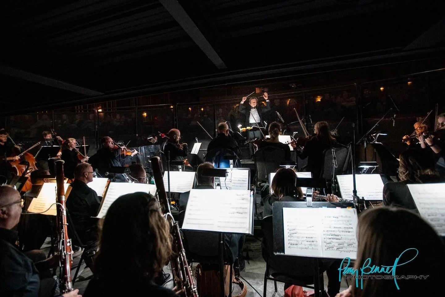 Orchestra performing in a dark concert hall with a conductor leading, and sheet music on music stands in front of musicians.