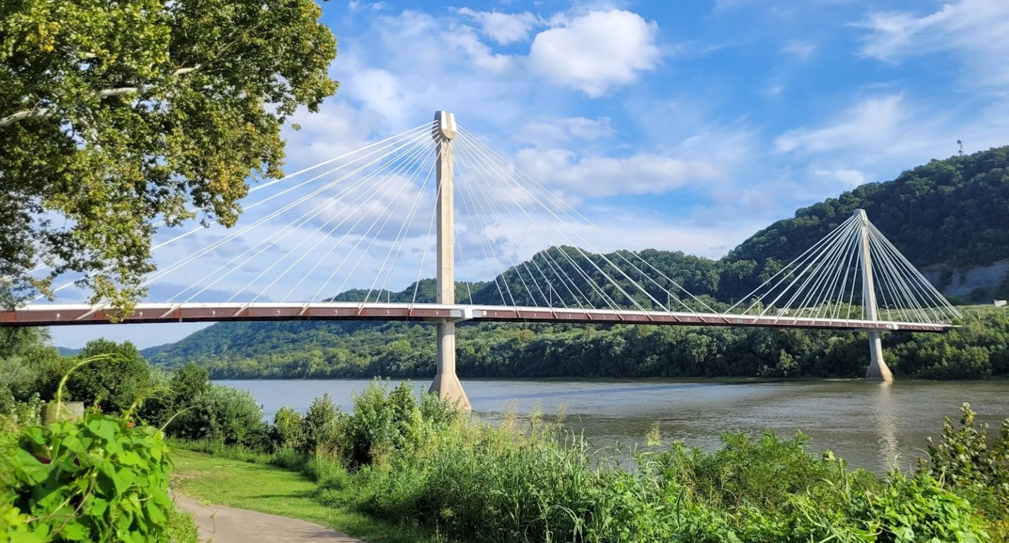 A modern suspension bridge over a river, surrounded by green trees and hills, under a partly cloudy sky.