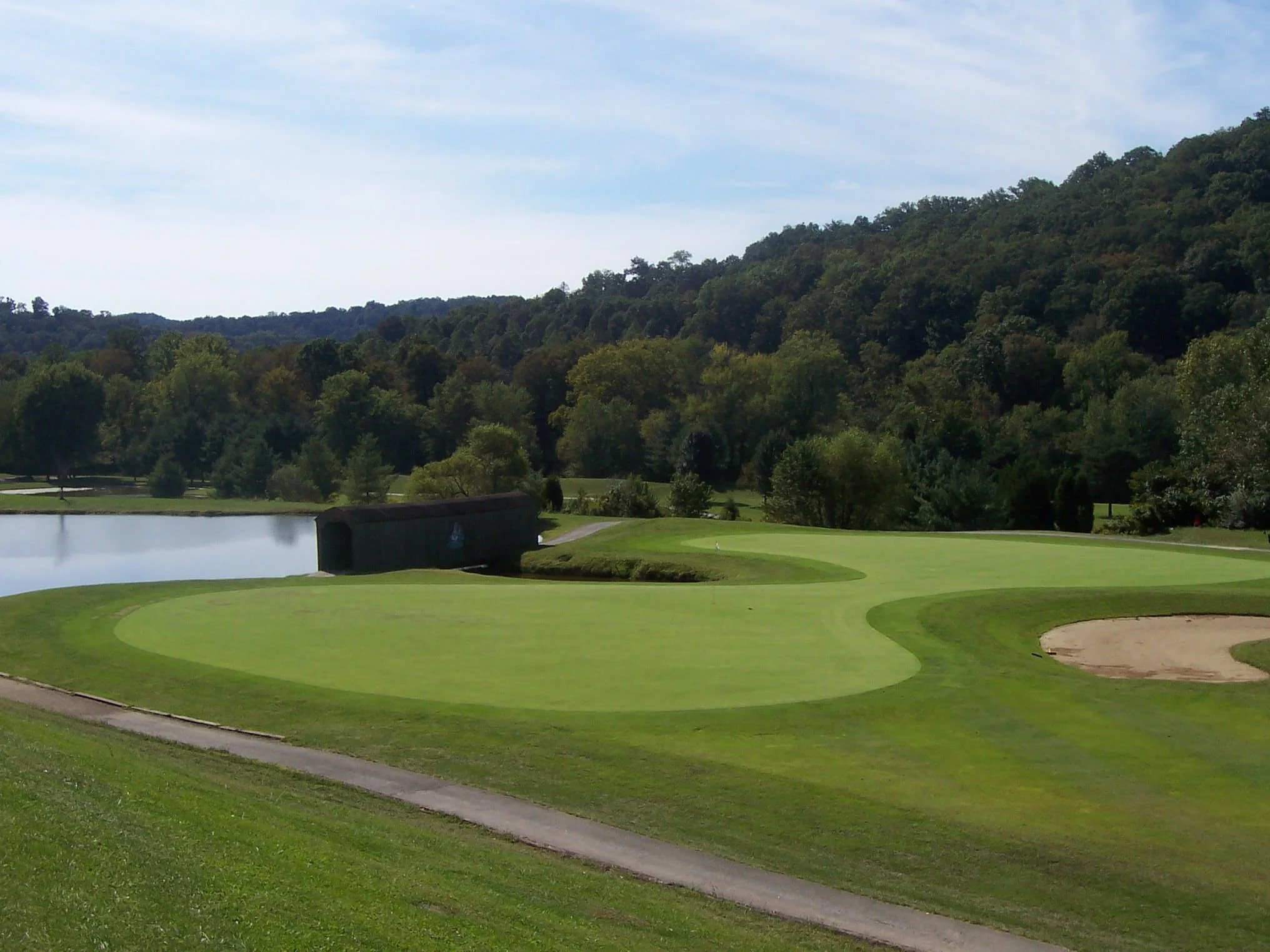 A golf course with a putting green, sand trap, and a water hazard, surrounded by trees and hills under a partly cloudy sky.