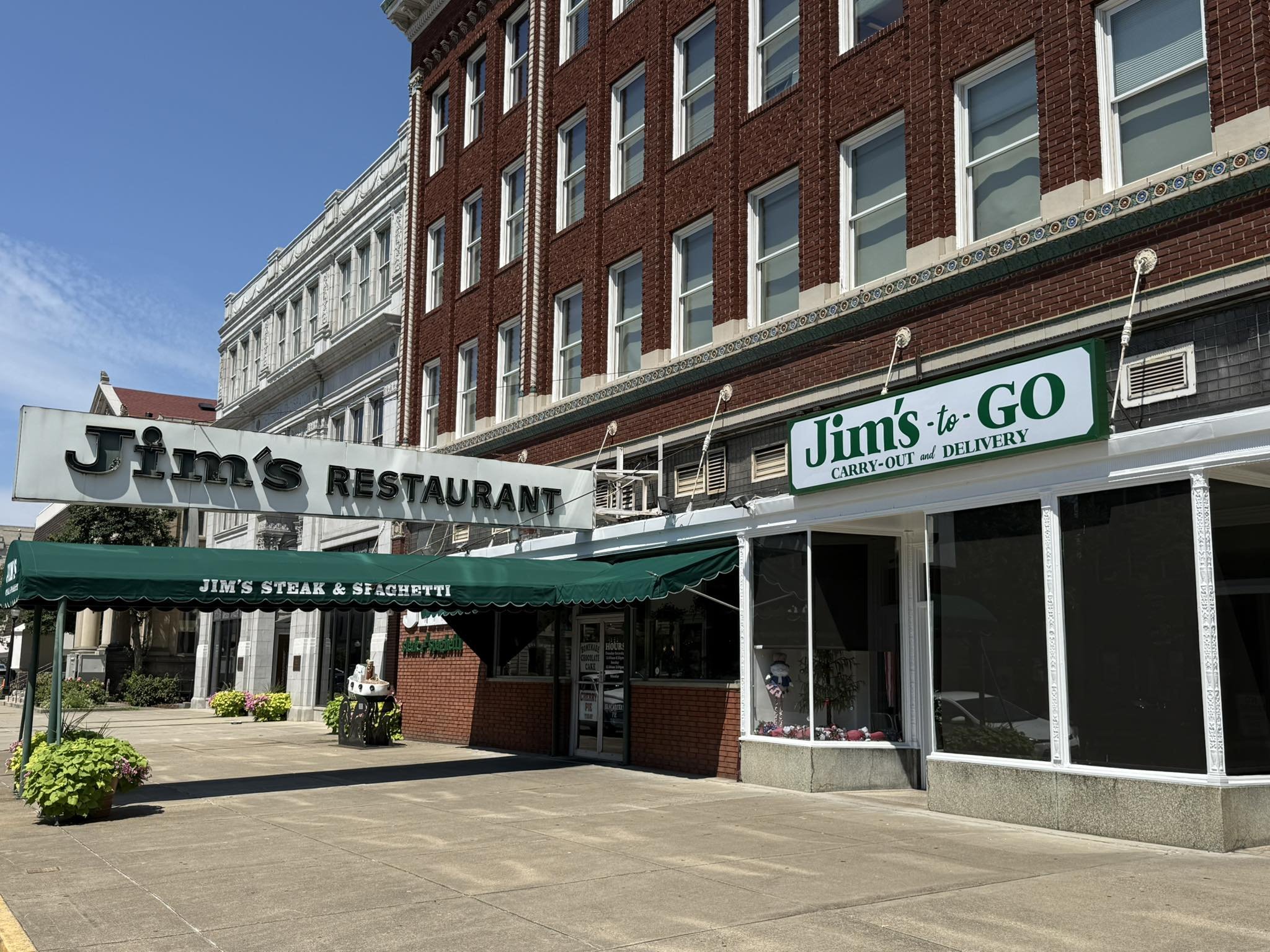 Exterior view of Jim's Restaurant and Jimmy's to-G carry-out and delivery restaurant located in a multi-story brick building, with a green awning over the entrance reading "Jim's Steak & Spaghetti," and signs indicating takeout and delivery services.